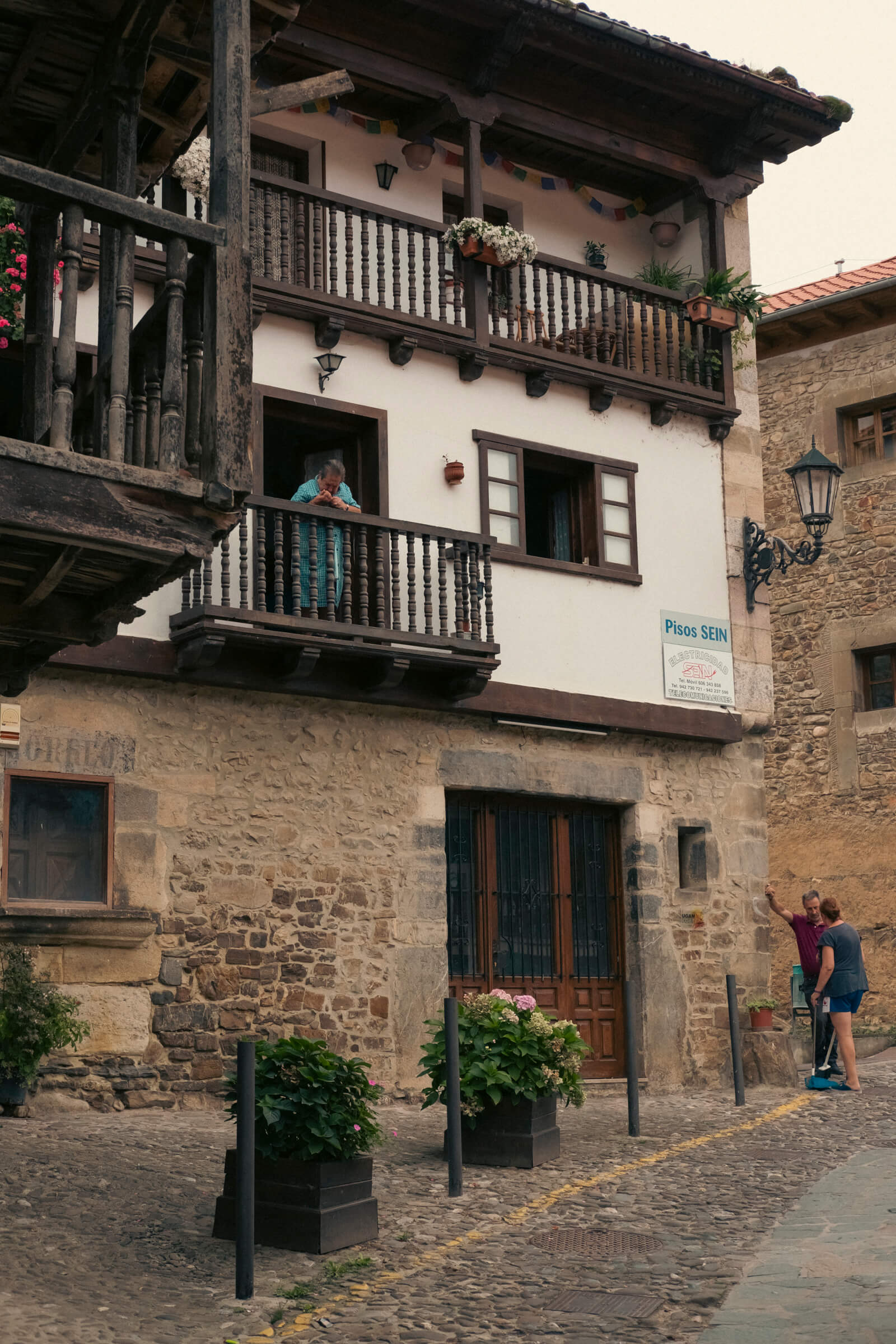 Escena de pueblo cántabro. Una casa típica montañesa de Cantabria con una persona asomada al balcón y dos abajo charlando en la calle