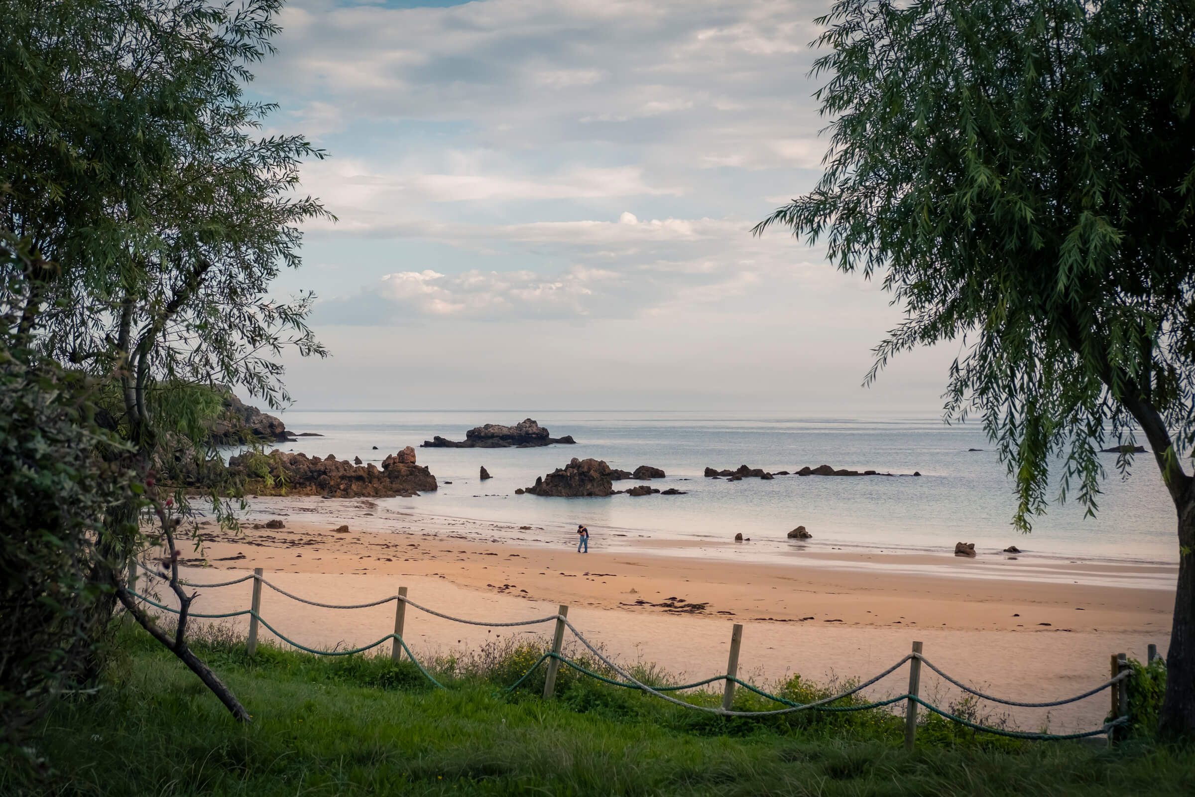 Pareja en un momento de intimidad aprovechando que no hay nadie en la Plaza de Toranda. Encuadrada por ramas de árboles a los lados se ve la playa al fondo y, muy de lejos, dos personas abrazándose y besándose