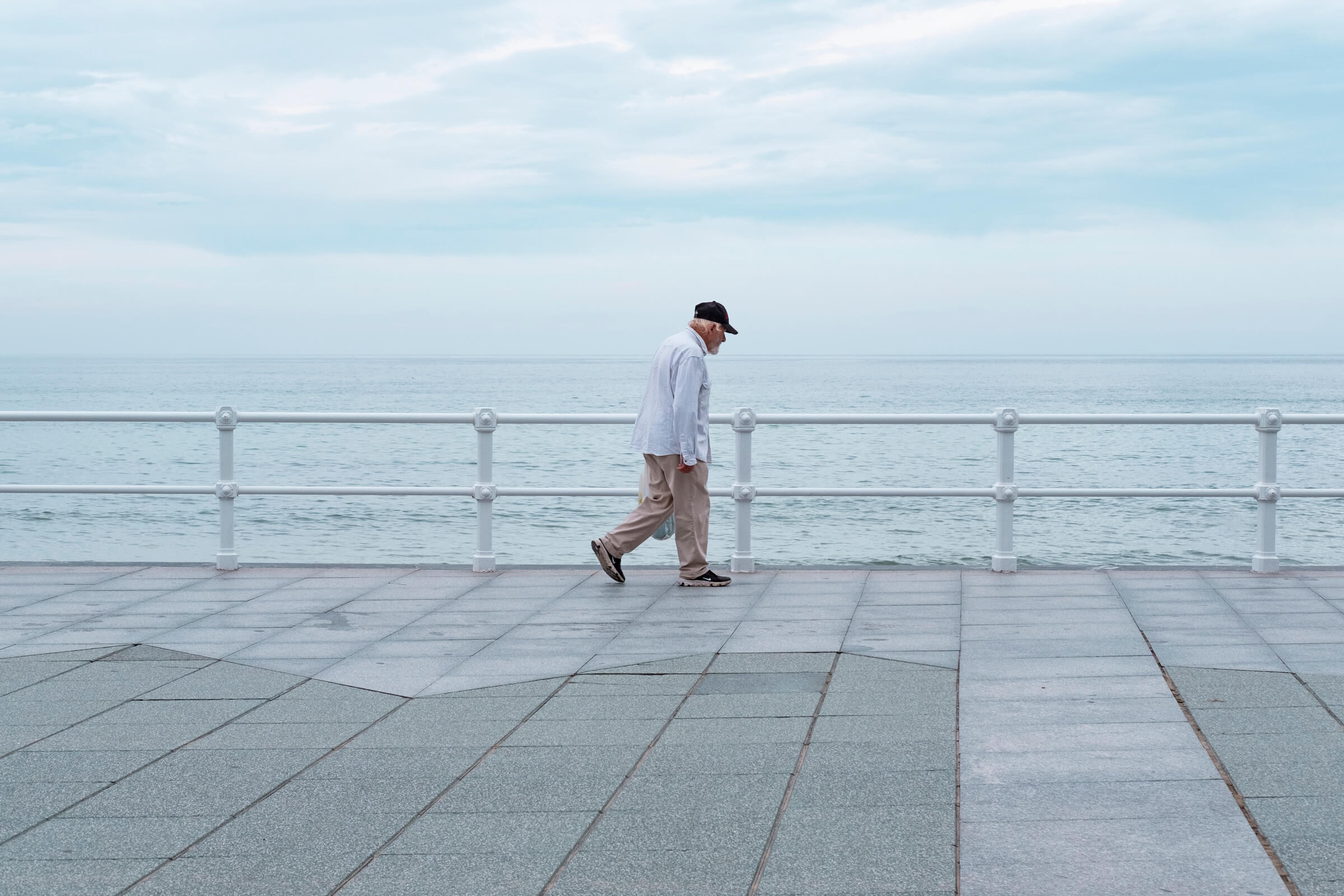 Paseo por el paseo. Señor caminando por el paseo de Gijón, se le ve avanzando de izquierda a derecha y encuadrado en el centro, con un color azul grisáceo en el cielo