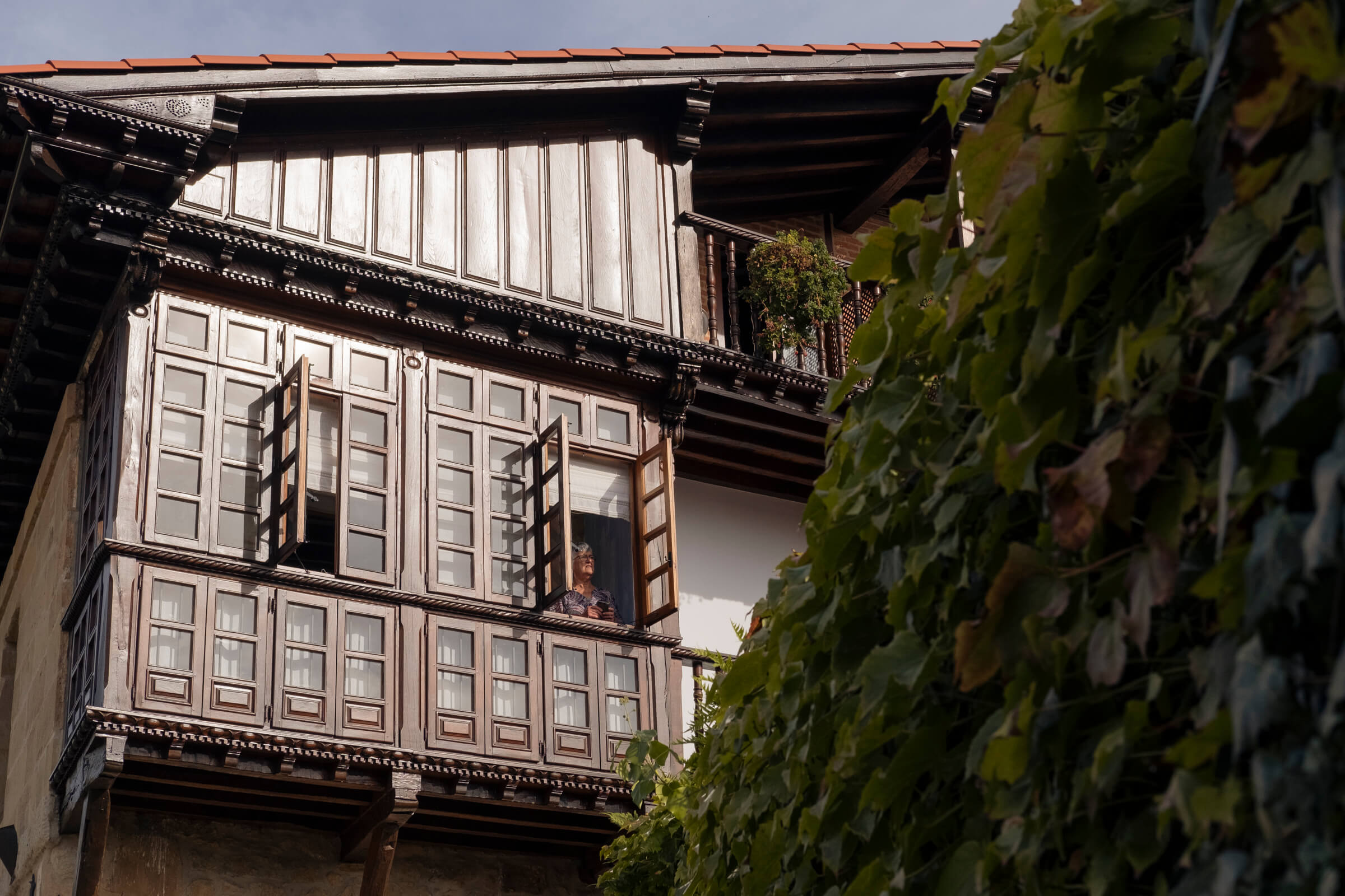 Un poquito de aire y sol por la mañanita. En una casa de madera se ven ventanas abiertas y una señora asomada al sol