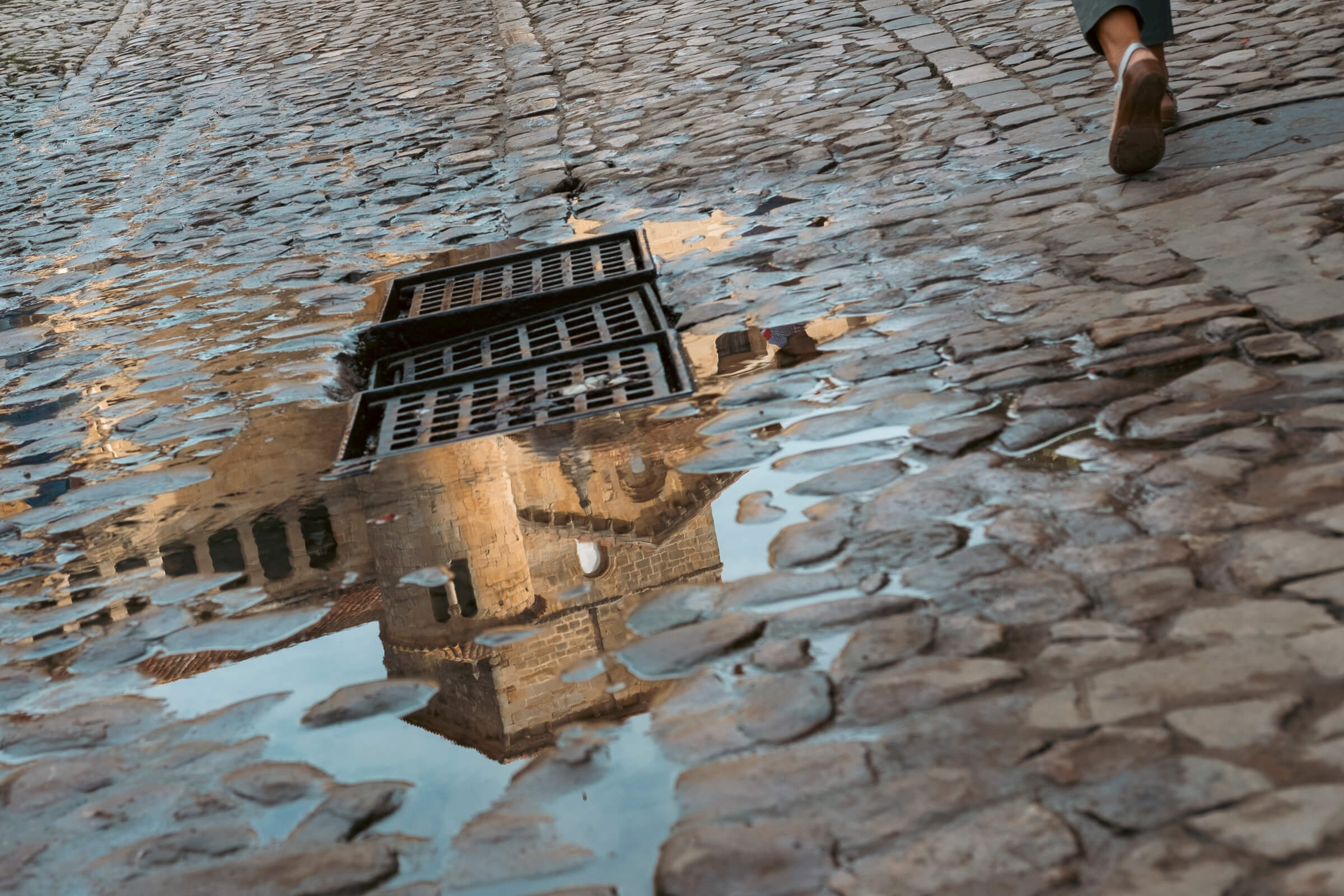La Colegiata de Santillana del Mar. Foto a un charco en el que se refleja parte del edificio antiguo y de la Colegiata de Santillana del Mar. En la esquina, se ve a una persona que pasa caminando al lado del charco
