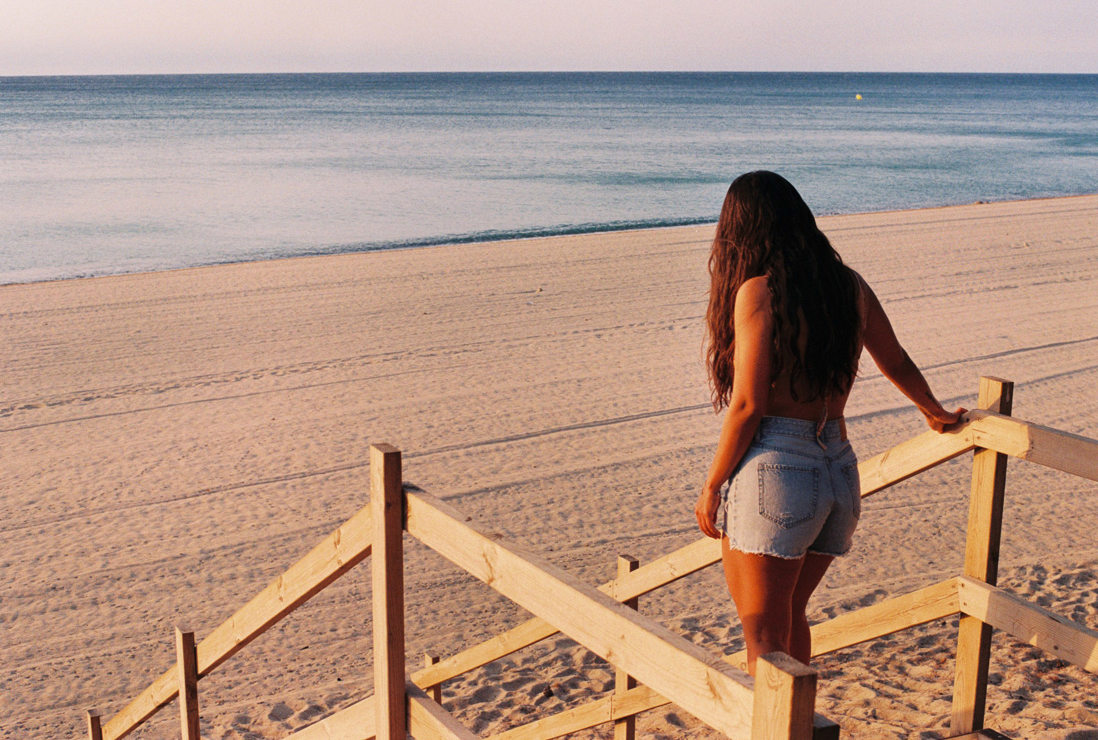 Fotografía de Laura bajando a la playa por una escalera de madera con el mar y la luz de amanecer de fondo, tomada con una cámara analógica de carrete