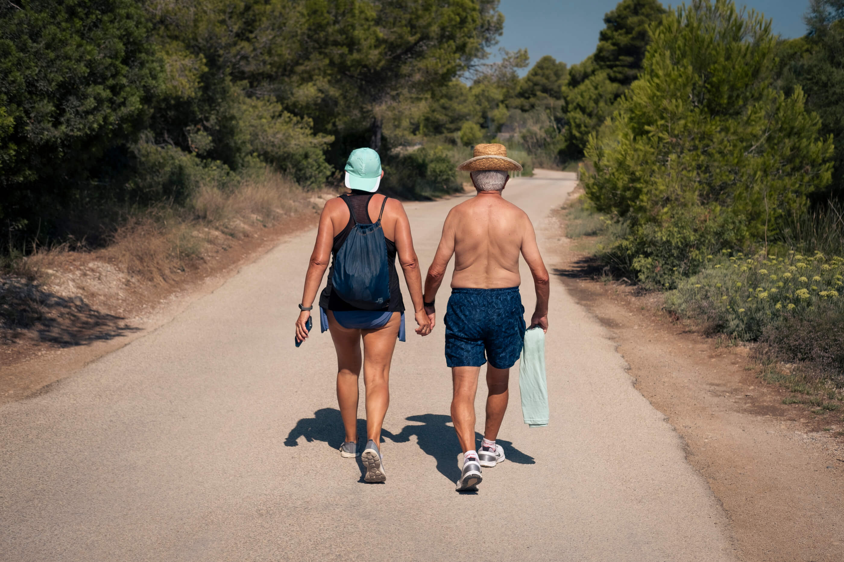 Mis padres paseando de la mano, cual novios jovenzanos. Mis padres de espaldas paseando de la mano con ropa veraniega por una carretera rural, con árboles a ambos lados de la misma