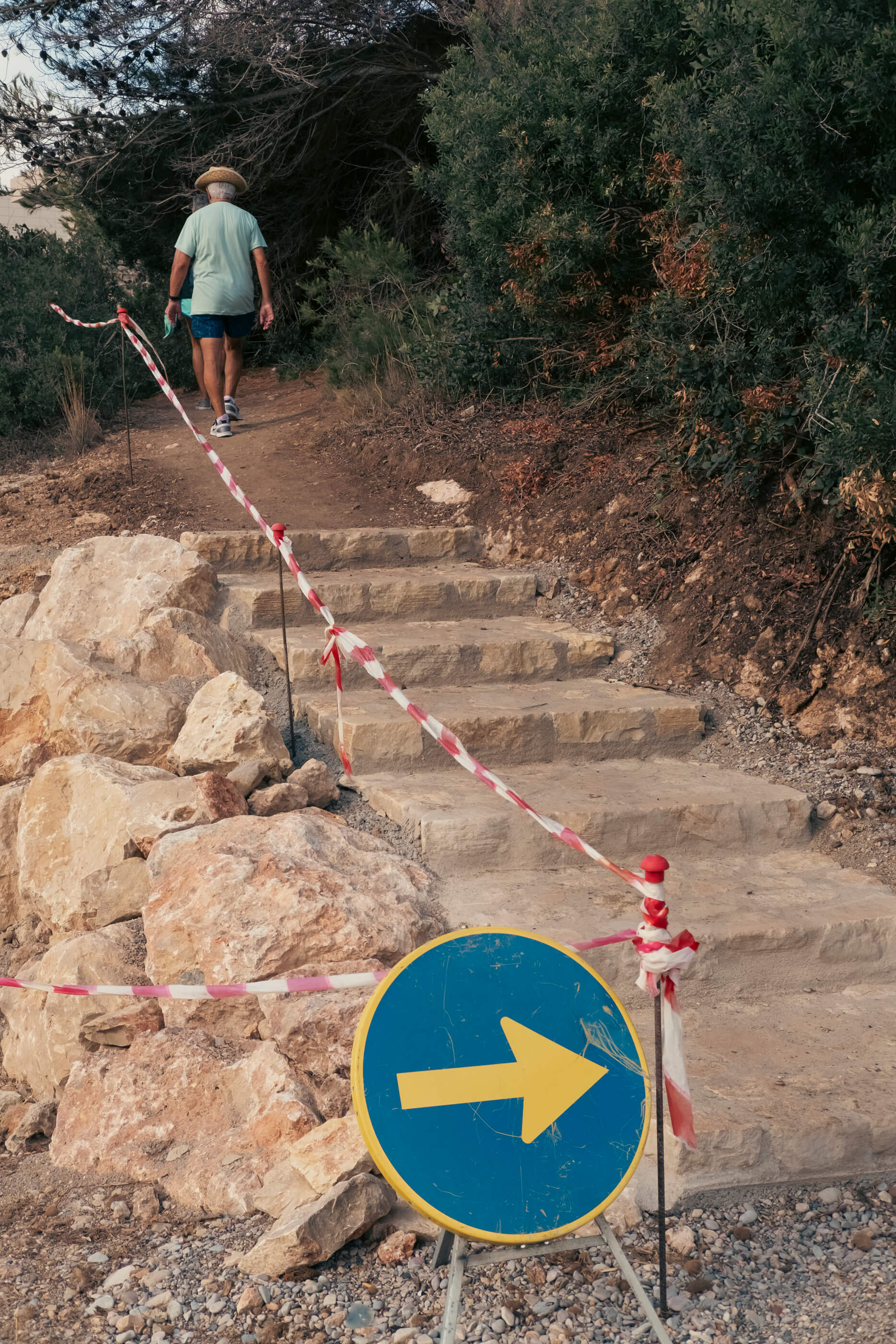 Escaleras en construcción en un sendero estrecho, con una señal y una banda indicando por dónde pasar, y alguien caminando al fondo