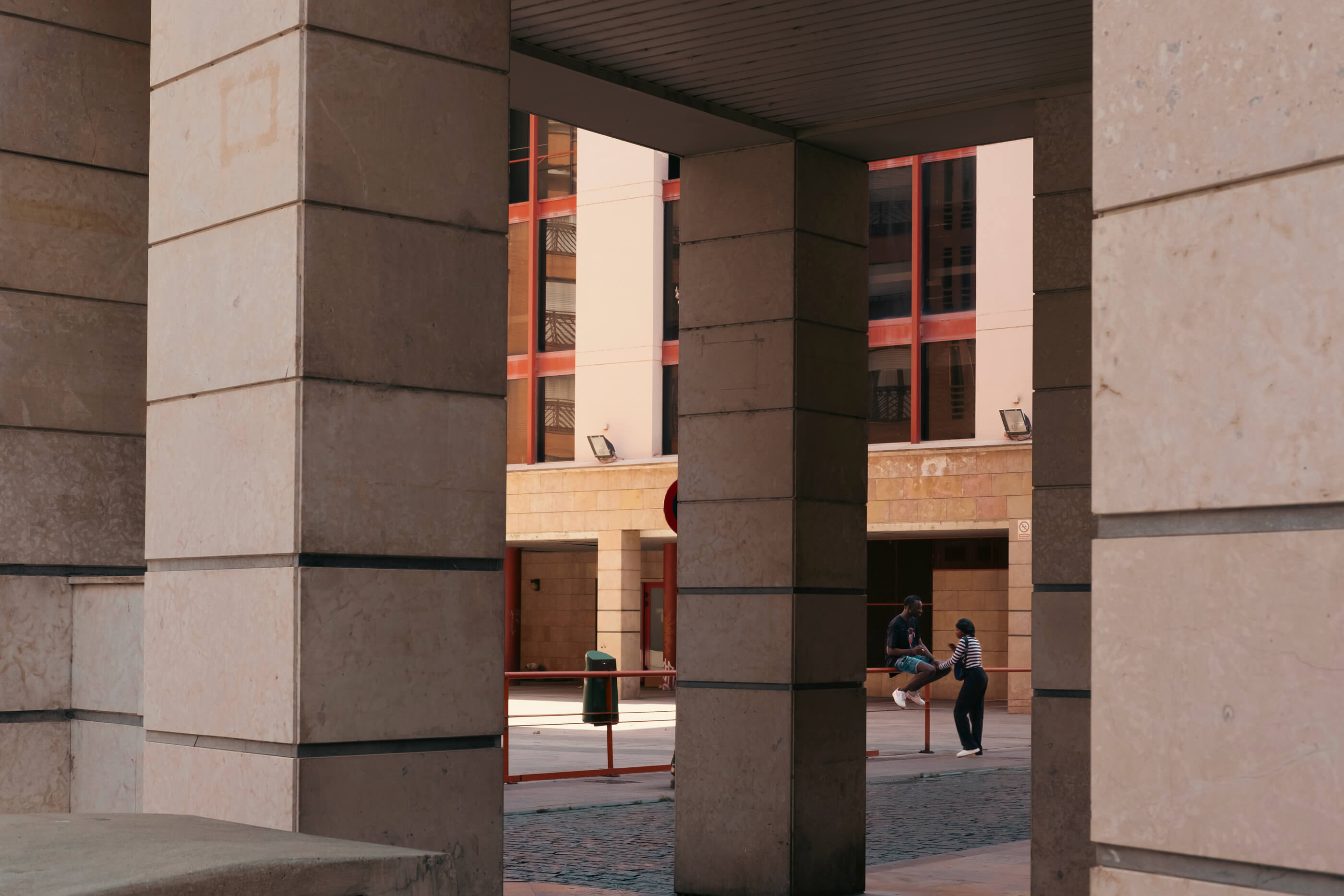 Un chico y una chica charlan en una plaza. El encuadre de la pareja juega con las perspectivas y las líneas rectas que forman las columnas y las ventanas
