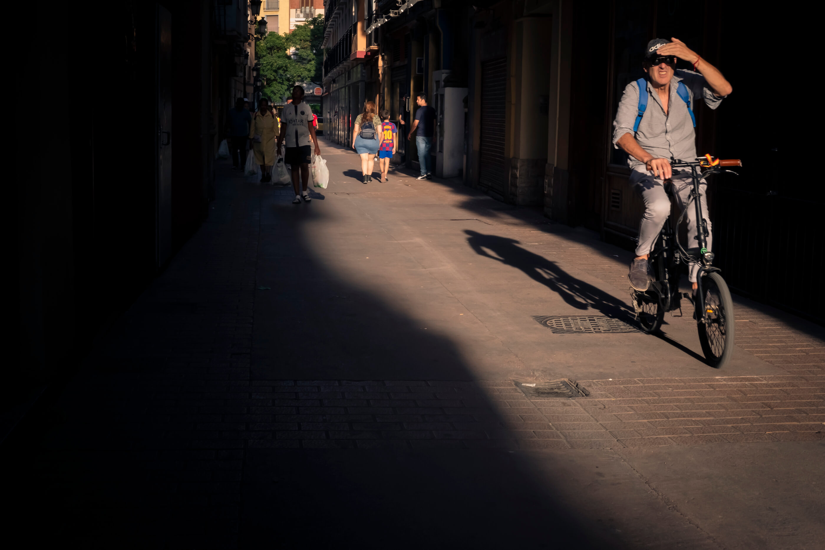 Juego de luces y sombras en la calle, con gente pasando por zonas oscuras, en sombra, y un ciclista por la zona iluminada y tapándose la cara porque le molesta el sol