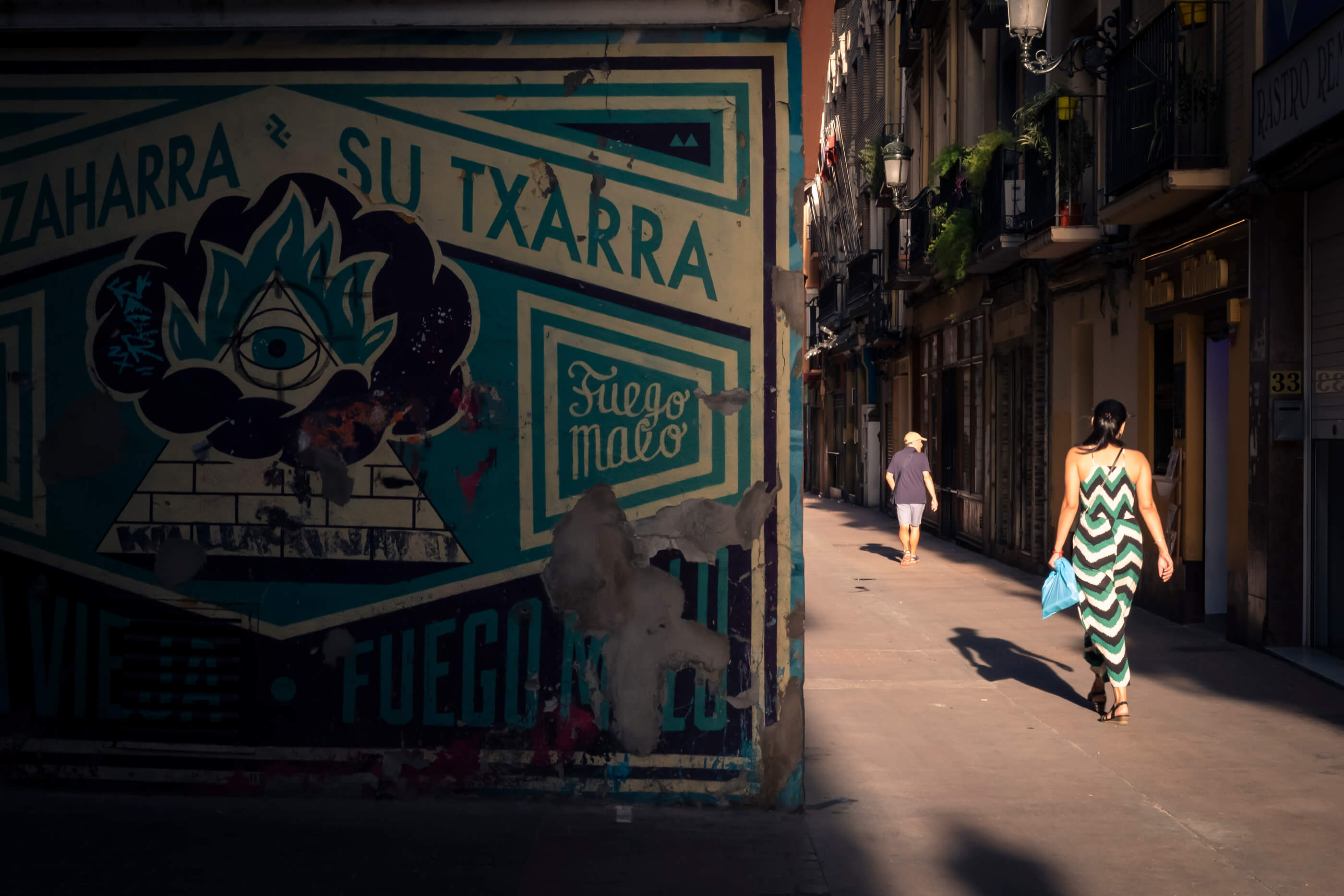 Foto callejera en la que se ve media foto con una pared que tiene un mural que coincide en colores y formas con el vestido de una chica que pasa en ese momento por la calle, justo al lado e iluminada por el sol