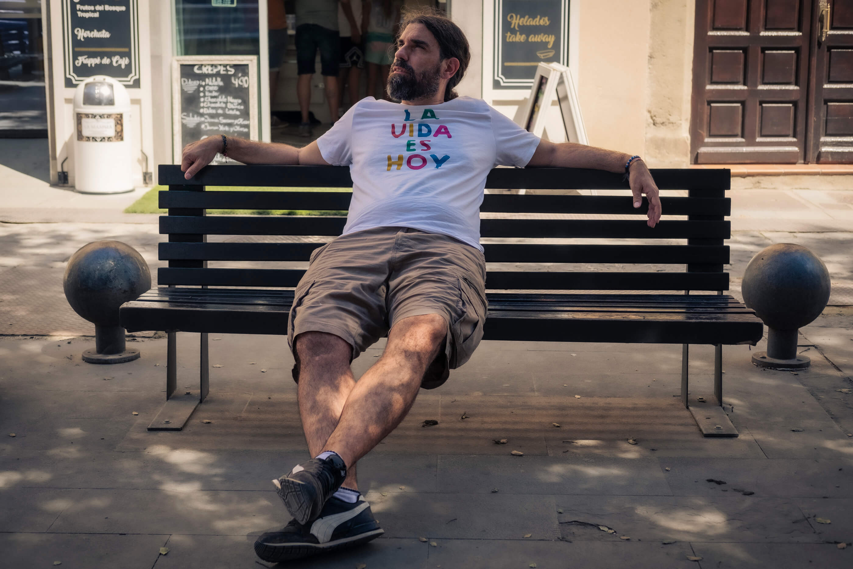 La vida es hoy. Chico sentado en u banco en la calle, con los brazos abiertos y ocupando todo el espacio, con una camiseta en la que se lee 'La vida es hoy'