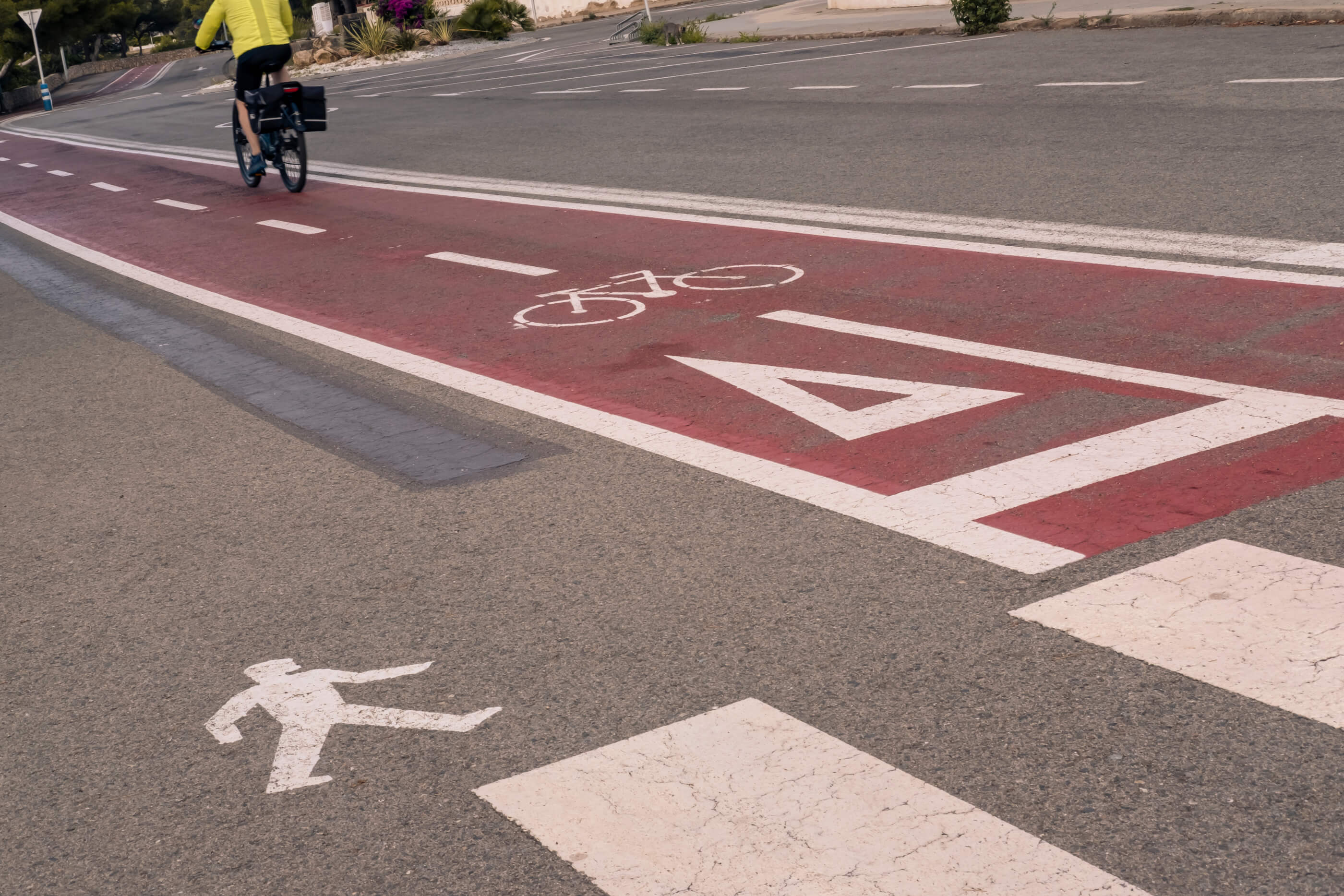 Juego de perspectiva con encuadre hacia el suelo, con un carril bici en diagonal, señalética en el suelo pintada de blanco y una persona pasando con su bici