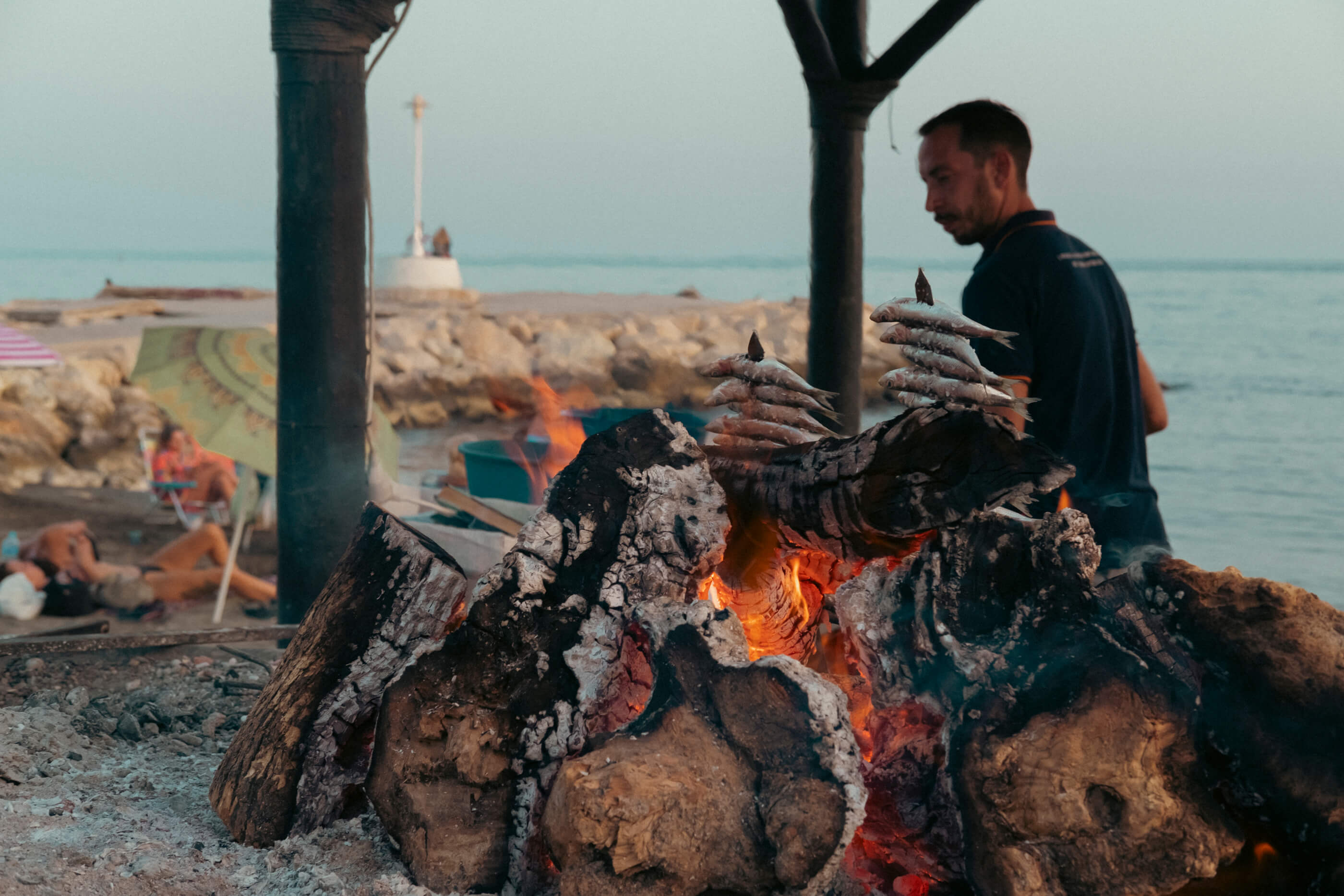 En primer plano el fuego vivo y los espetos de sardinas típicos de Málaga, de fondo la playa con gente aprovechando las últimas horas de sol