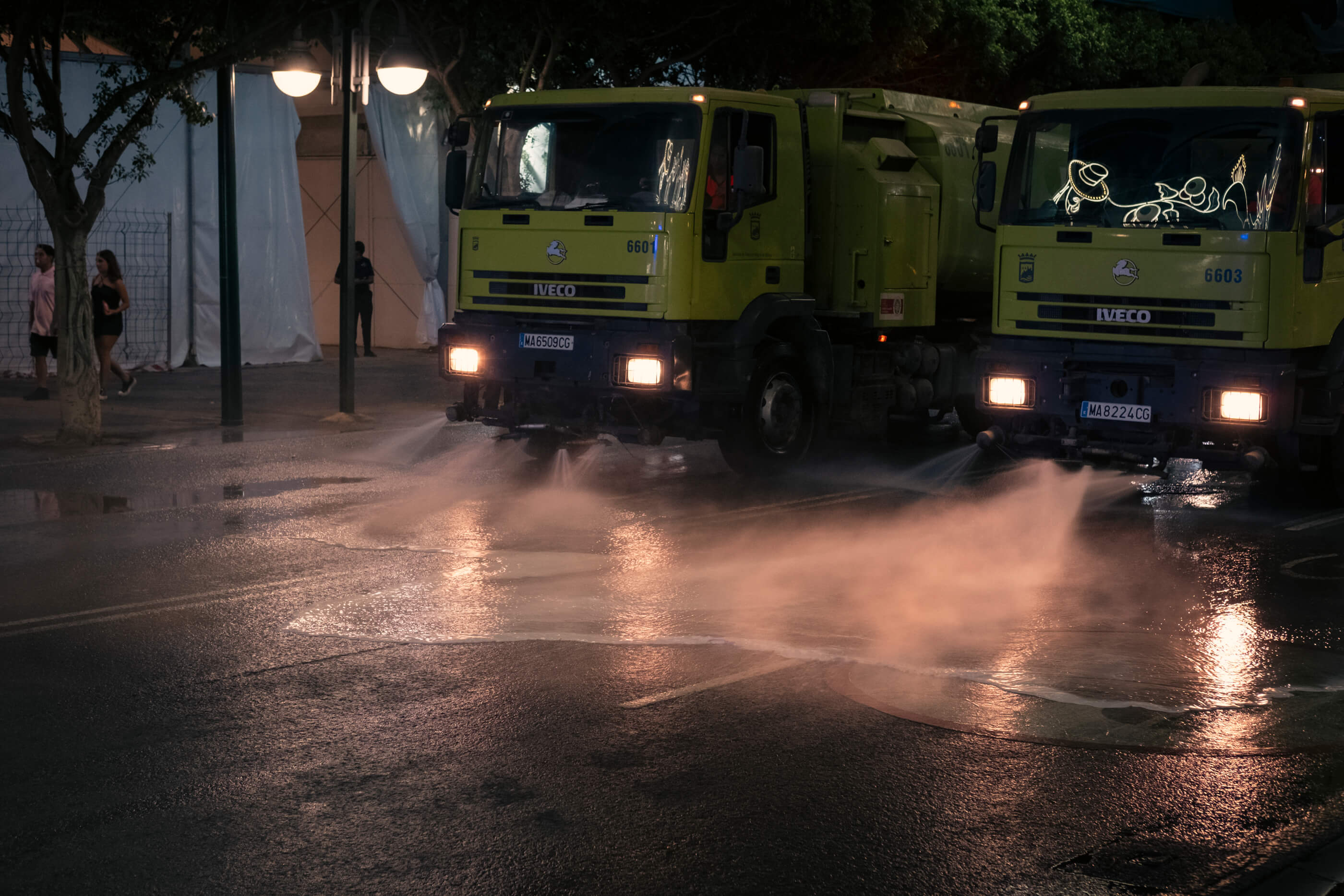 Camiones de limpieza por la noche en La Feria de Málaga. Foto nocturna con la iluminación de los faros de los camiones de limpieza que avanzan regando la calzada de una de las calles en La Feria de Málaga