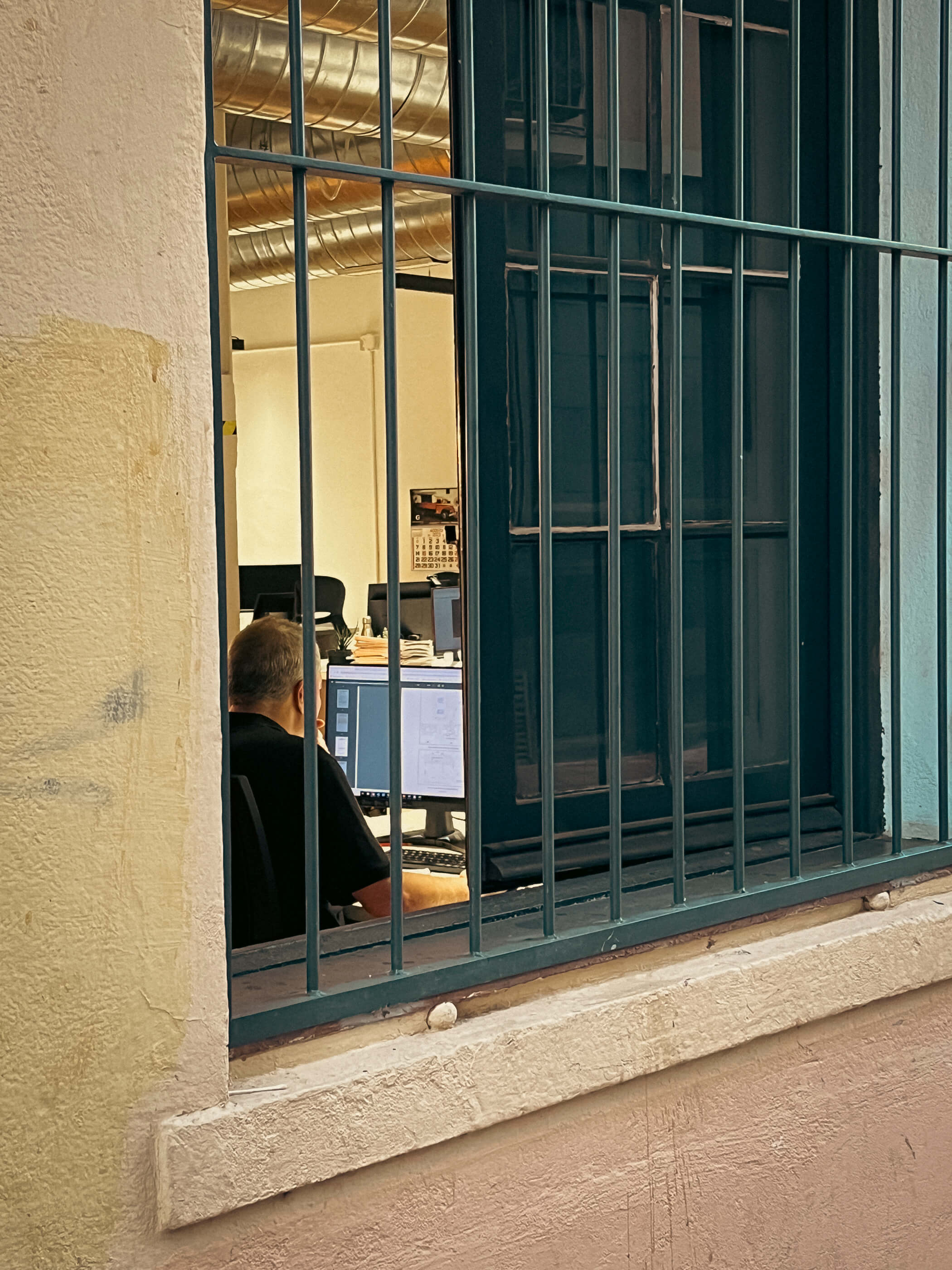 Cárceles modernas. Una persona trabajando en la oficina. La composición destaca la ventana con barrotes en primer plano y deja ver la persona trabajando dentro