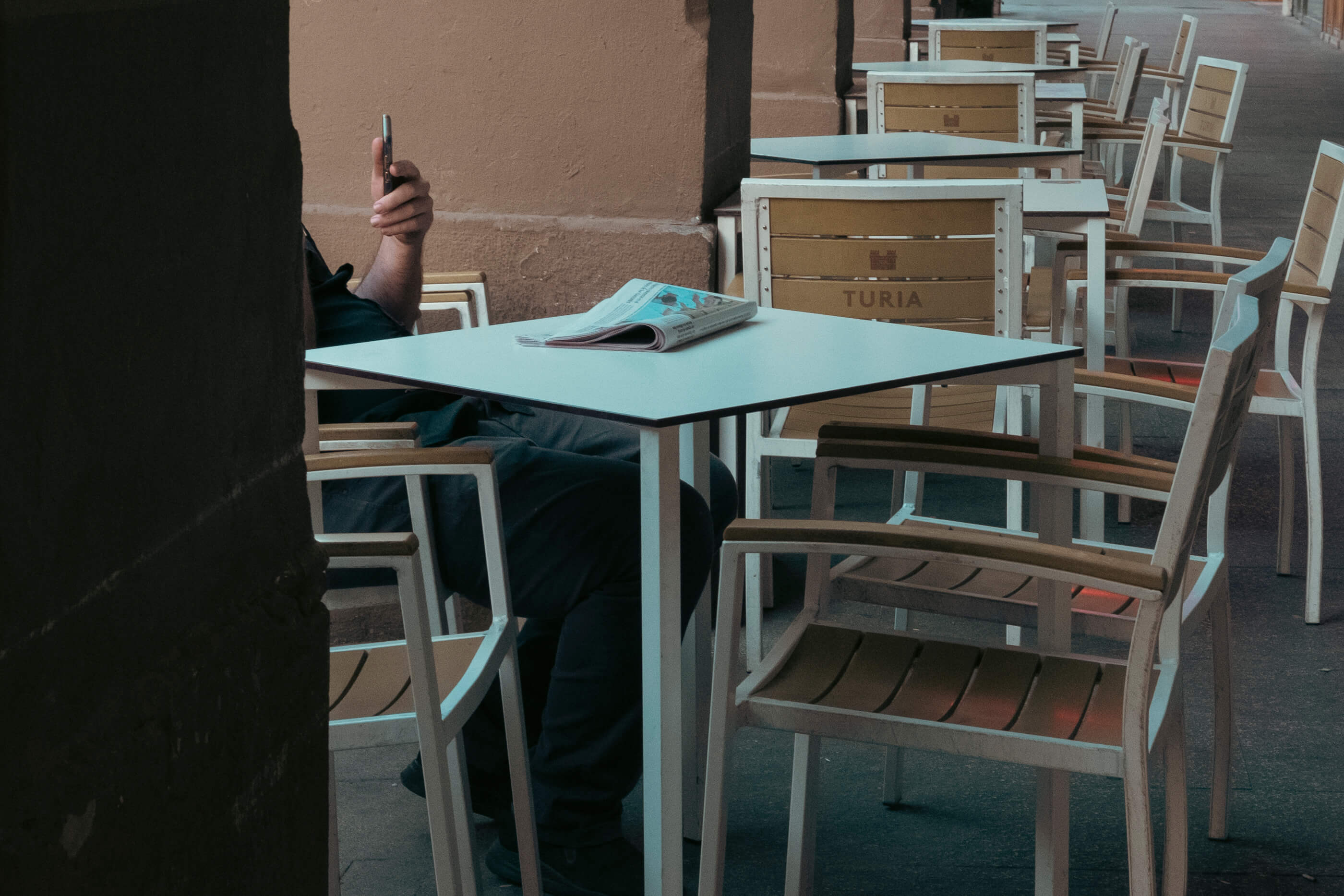 Revisando la actualidad por la mañana. Una terraza con todas las meses vacías salvo una en la que hay un señor mirando el móvil y con el periódico en la mesa. El encuadre hace que la columna tape al señor y solo se ve su mano con el móvil y sus piernas