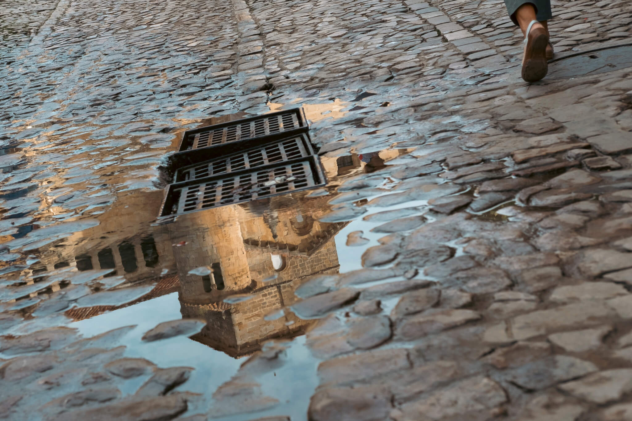 La Colegiata de Santillana del Mar. Foto a un charco en el que se refleja parte del edificio antiguo y de la Colegiata de Santillana del Mar. En la esquina, se ve a una persona que pasa caminando al lado del charco
