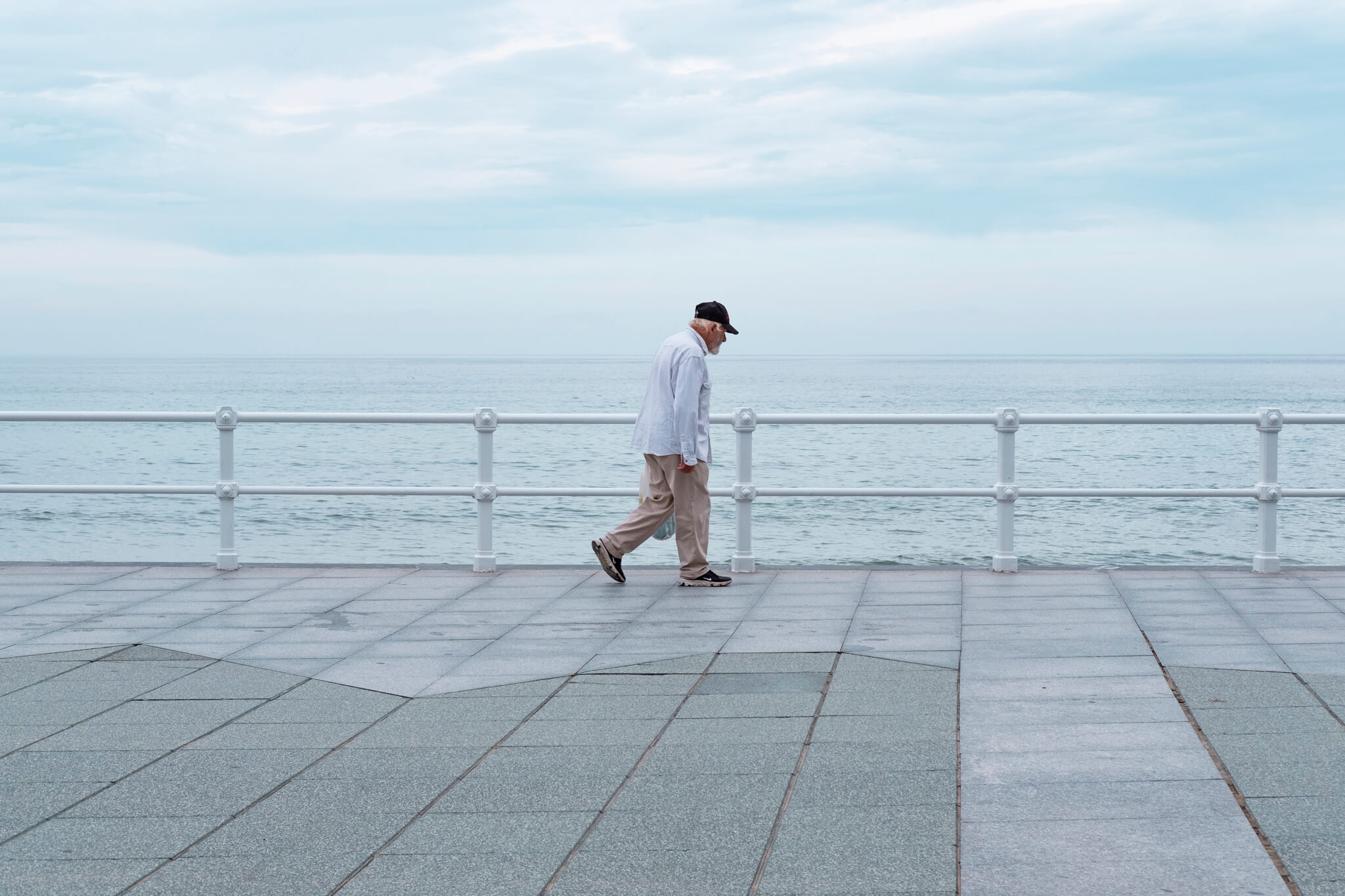 Paseo por el paseo. Señor caminando por el paseo de Gijón, se le ve avanzando de izquierda a derecha y encuadrado en el centro, con un color azul grisáceo en el cielo