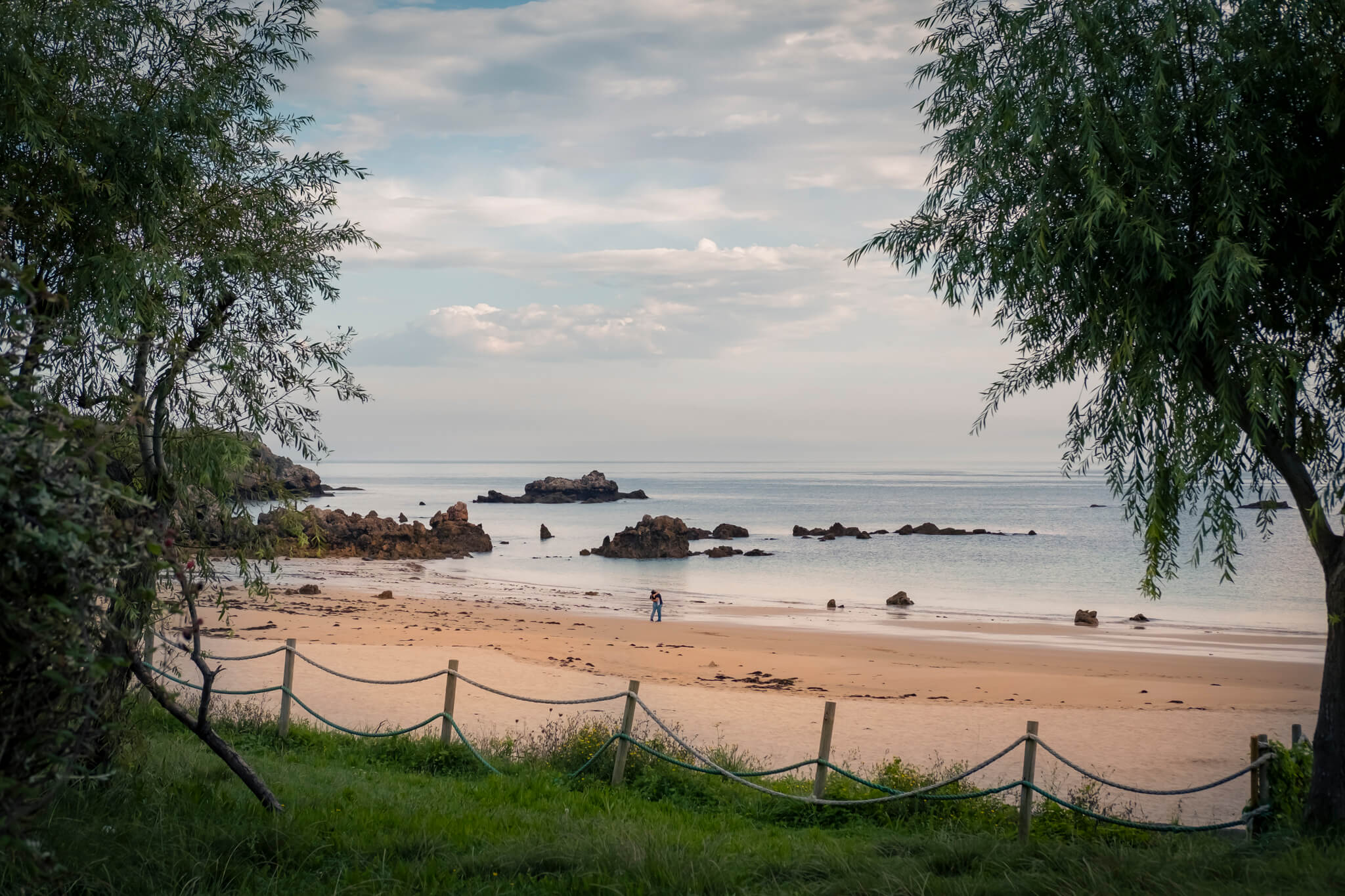 Pareja en un momento de intimidad aprovechando que no hay nadie en la Plaza de Toranda. Encuadrada por ramas de árboles a los lados se ve la playa al fondo y, muy de lejos, dos personas abrazándose y besándose