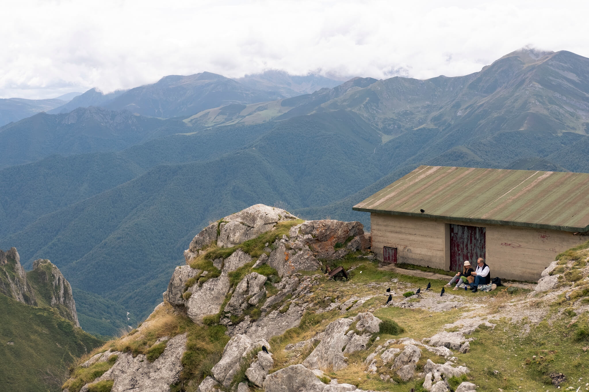 Almuerzo con invitados. Y con vistas. Arriba en Fuente Dé, una pareja comiendo algo sentados detrás del refugio, con varios pájaros pidiéndoles comida y con el valle de fondo