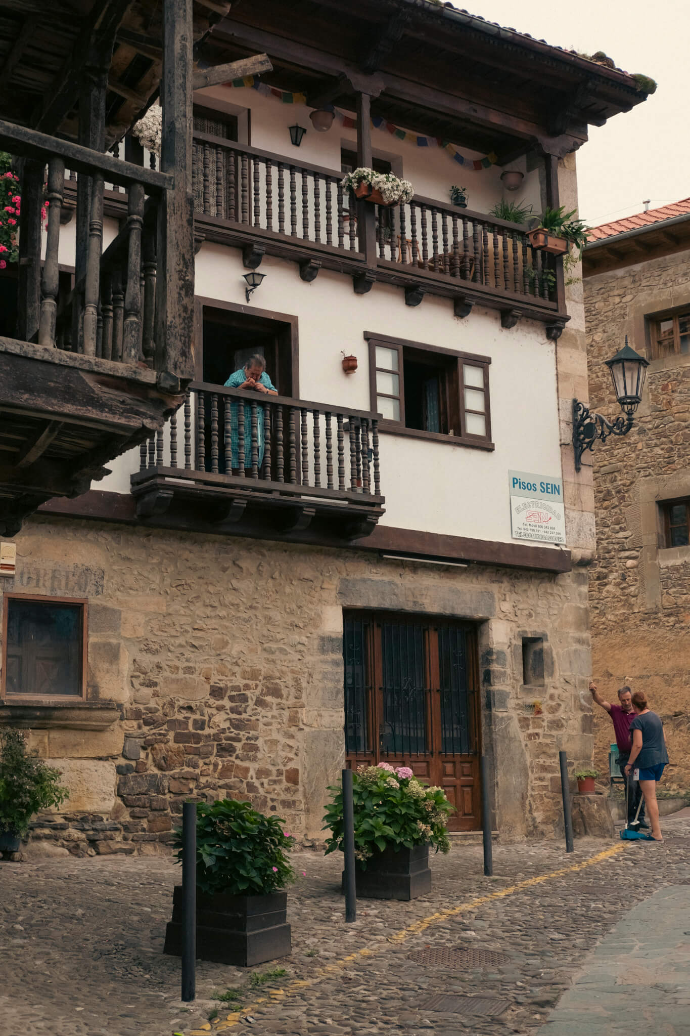 Escena de pueblo cántabro. Una casa típica montañesa de Cantabria con una persona asomada al balcón y dos abajo charlando en la calle