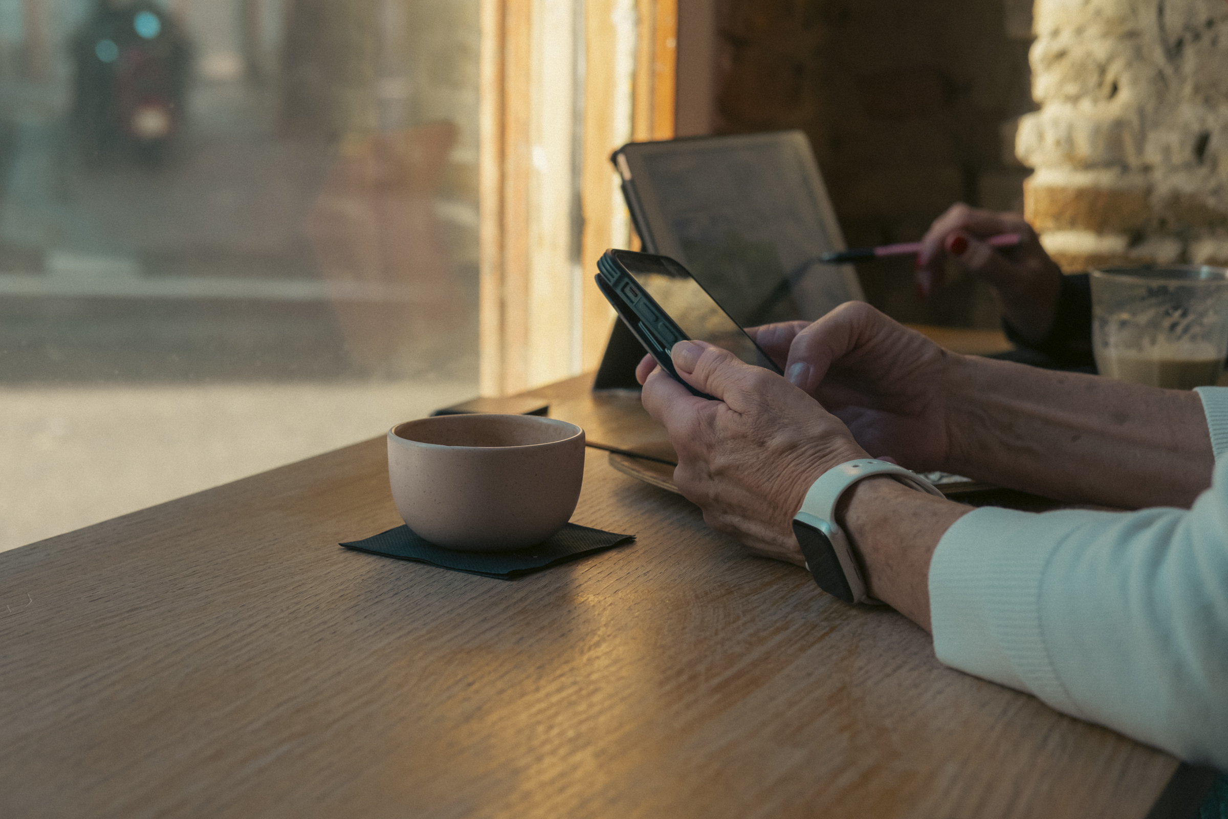 Dos señoras trabajando en un café. Se ve la mesa de un café, pegada a una ventana soleada, y el encuadre muestra las manos de dos personas, una sujetando un móvil y la otra con una tablet