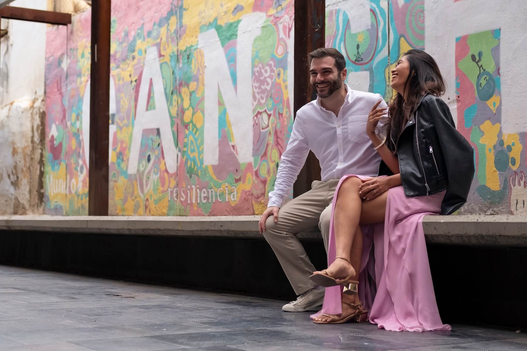 Mis amigos Héctor y Kitt, riéndose, sentados en un banco de cemento un mural muy colorido en la pared de detrás