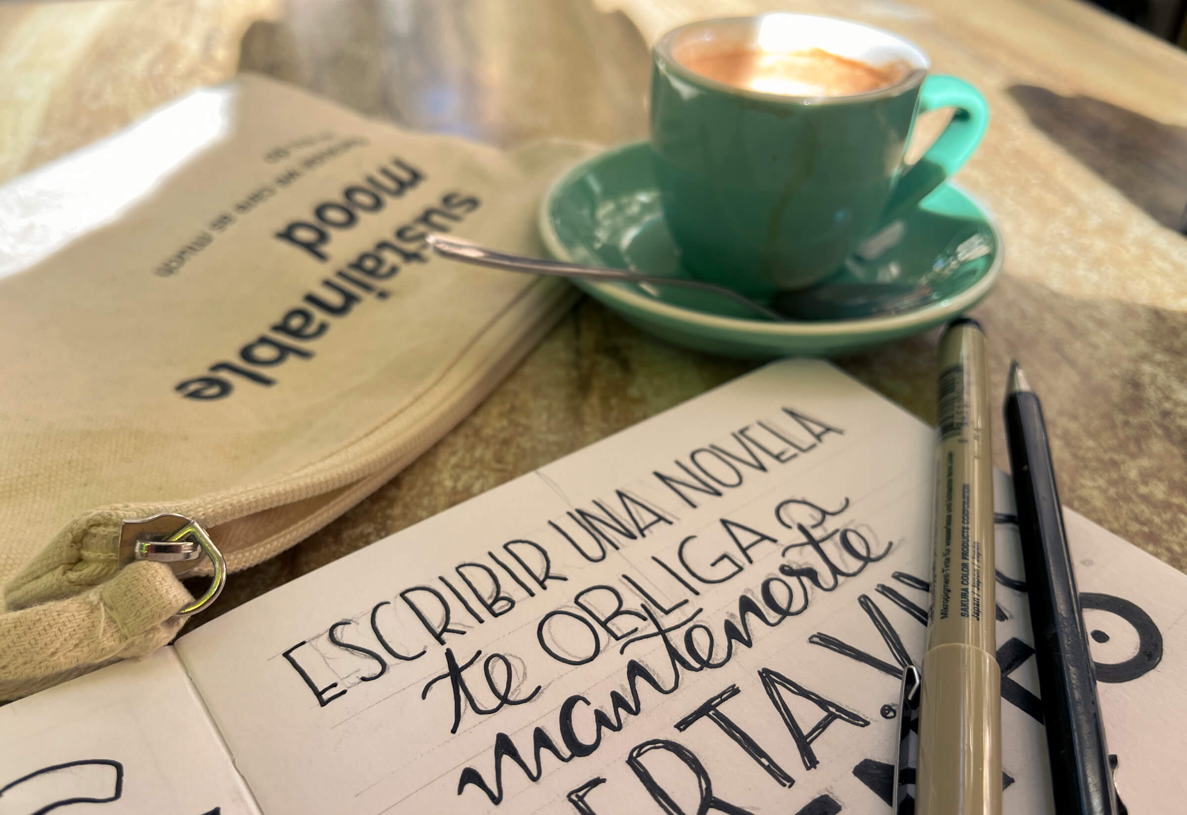 Foto del proceso, escribiendo la cita en el cuaderno. Foto del cuaderno sobre la mesa, con la cita a medio escribir, los rotuladores y una taza de café en una mesa soleada de una cafetería