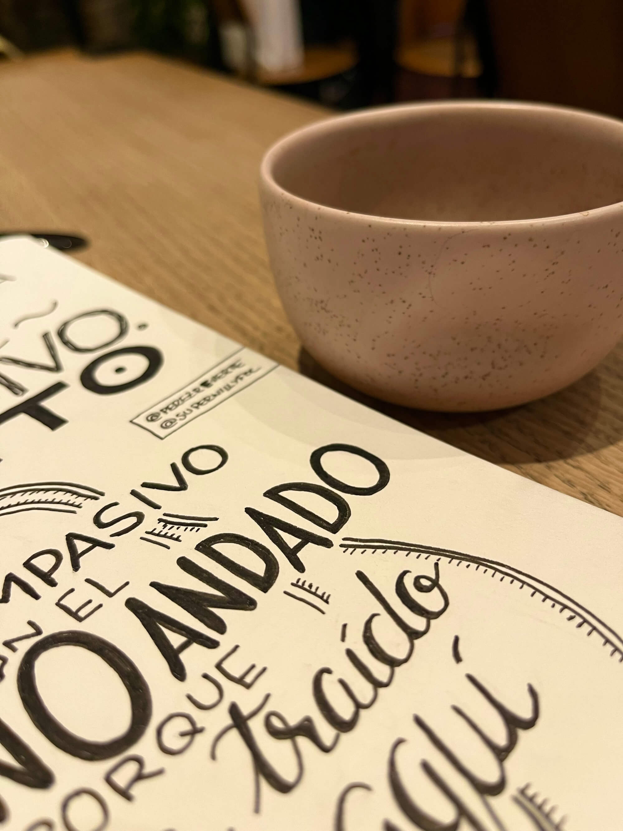 Foto del proceso, escribiendo la cita en el cuaderno. Foto del cuaderno sobre la mesa, con la cita a medio escribir, los rotuladores y una taza de café en una mesa soleada de una cafetería