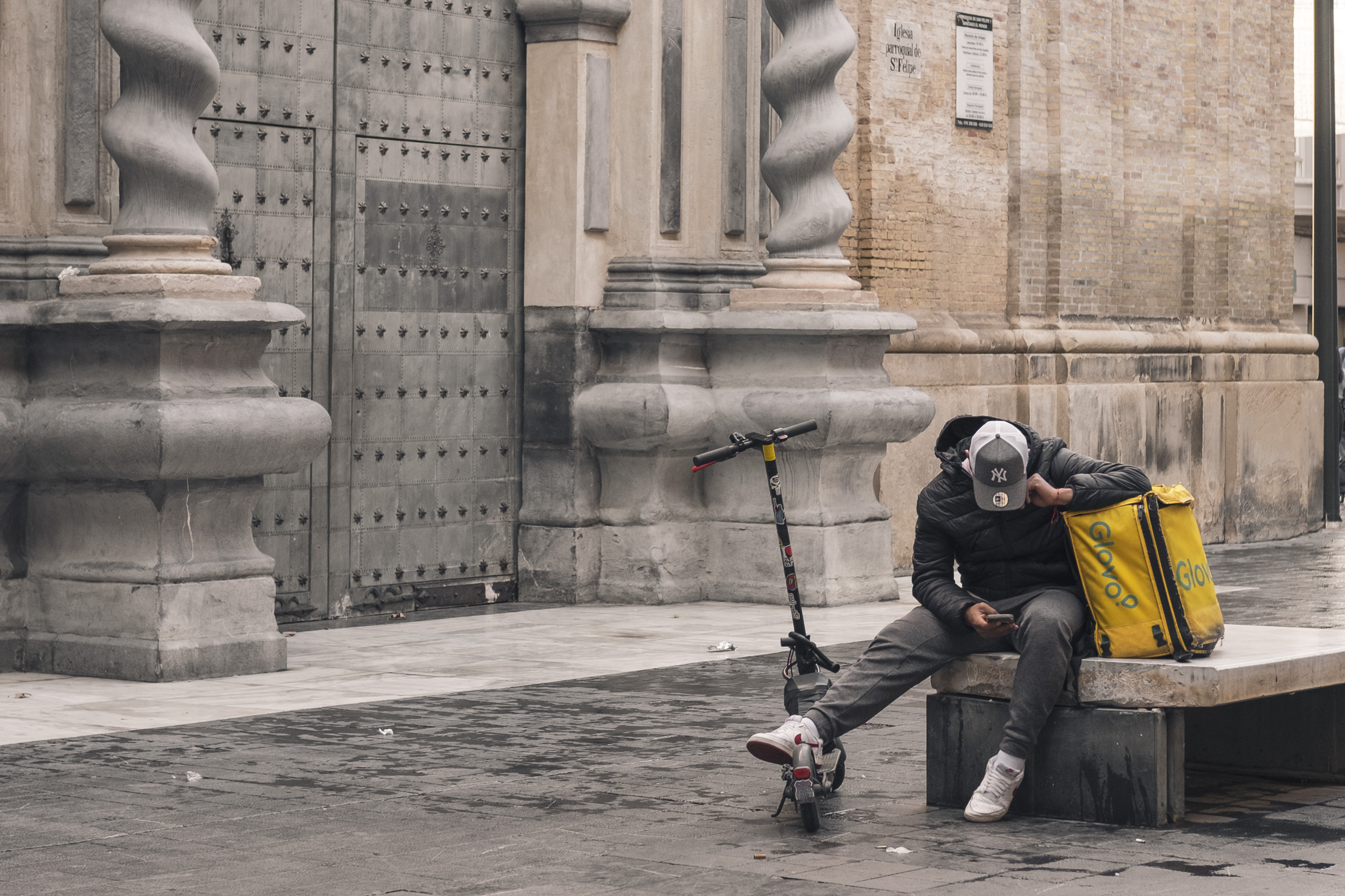 Un repartidor de Glovo espera en un banco, delante de una iglesia, mirando el móvil