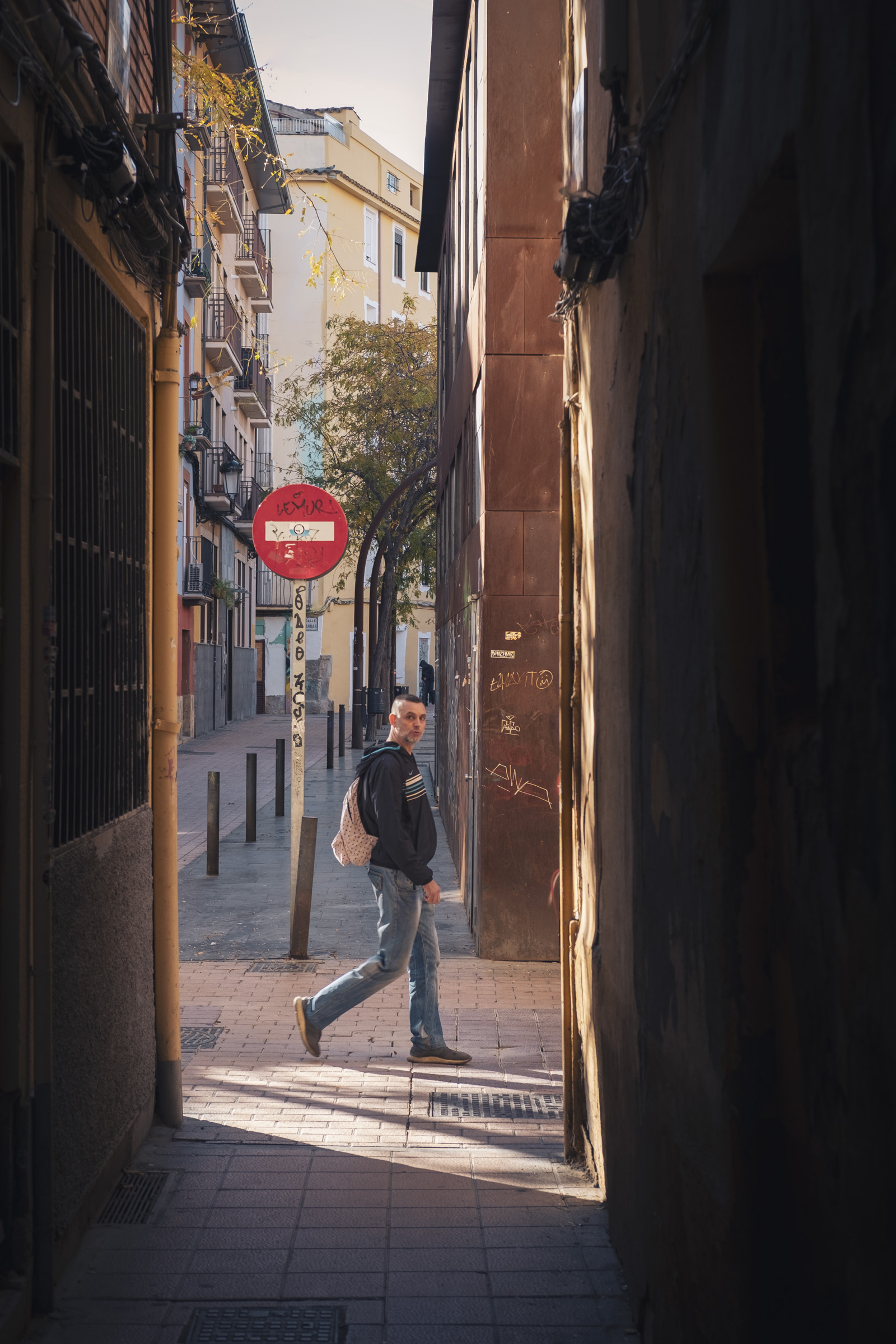 Foto encuadrada desde una calle muy estrecha y, al final de la misma, en el cruce con la siguiente calle, un chico pasa justamente en ese momento y mira a la cámara