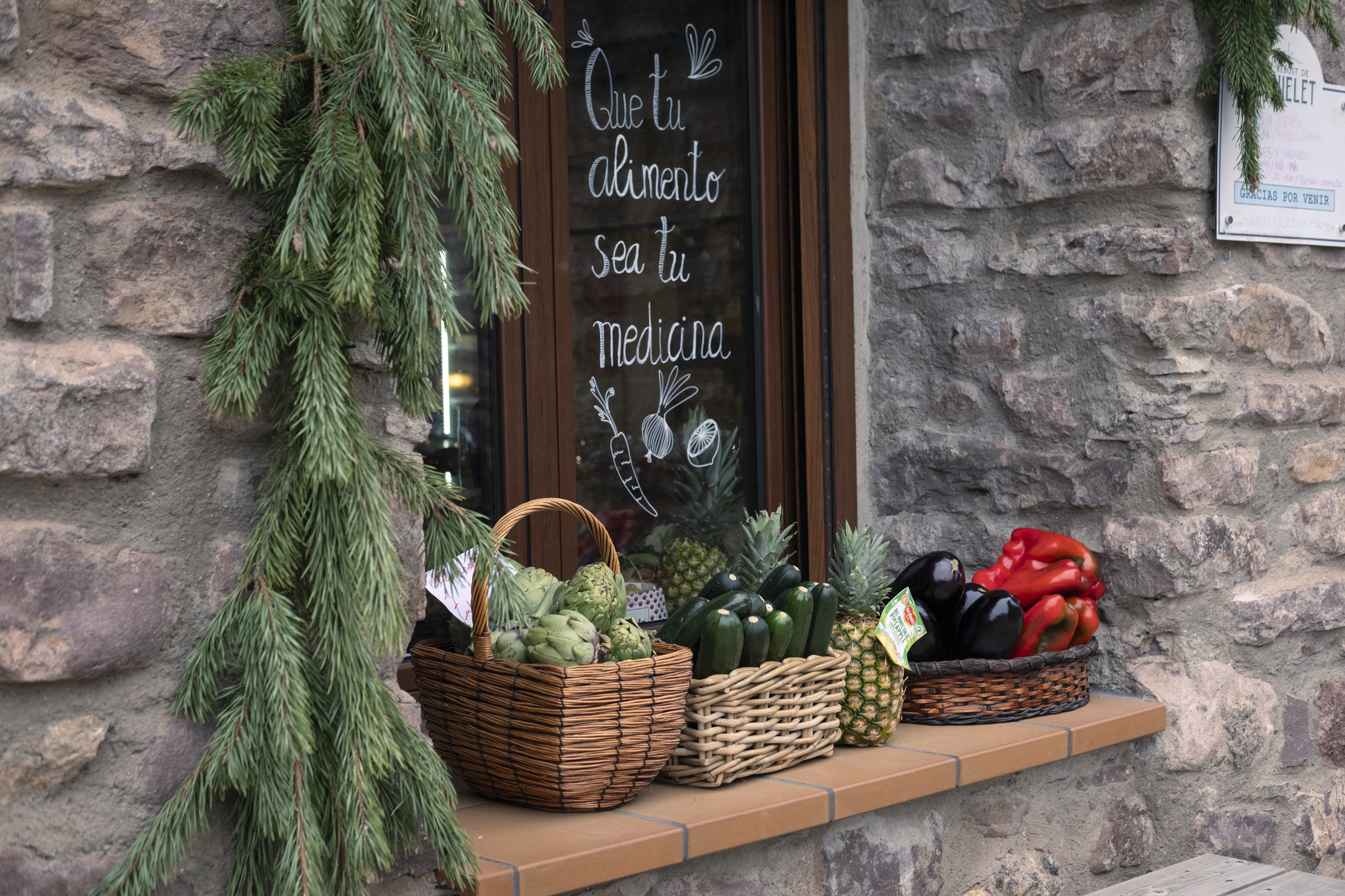 Tiendas pirenaicas que no necesitan refrigeración. Una tienda de frutas y verduras pone parte del género en la ventana para que se vea bien la calidad y se lee un cartel en el cristal que dice "Que tu alimento sea tu medicina"