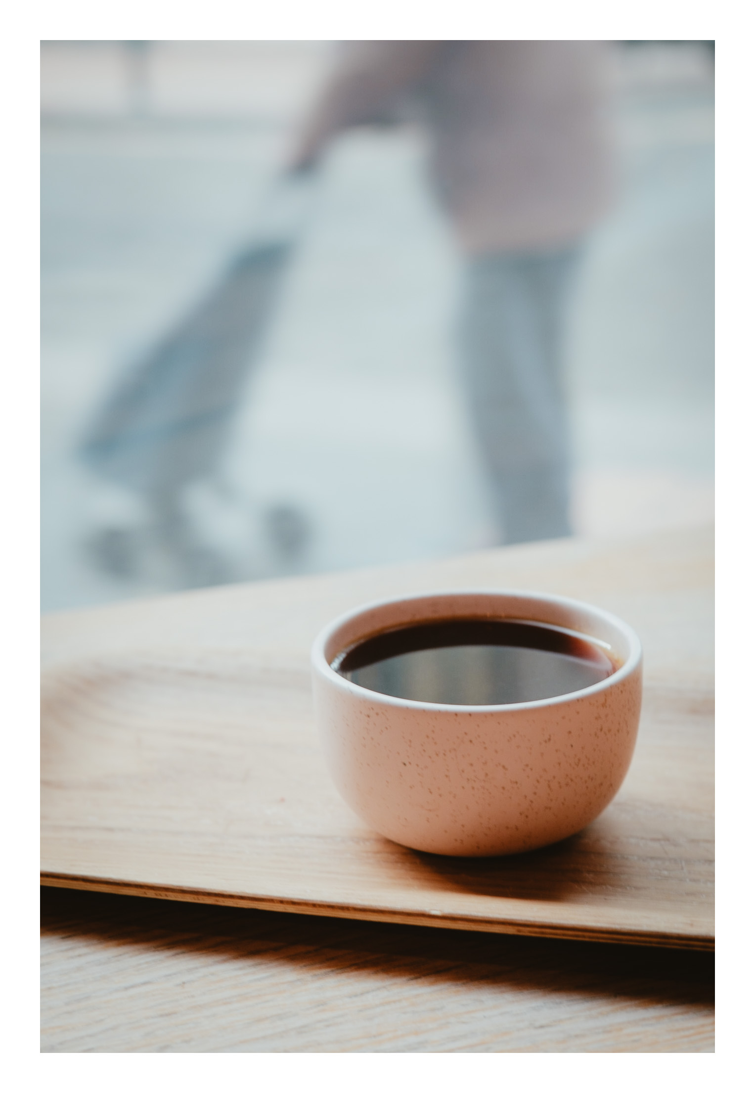 Placer I: un buen café especialidad en una taza bonita, mesa de madera y cristalera enorme a pie de calle. Justicia & Coffee. Taza de café en primer plano y, de fondo, una cristalera que da a la calle deja entrever a alguien pasando caminando con un carro de la compra