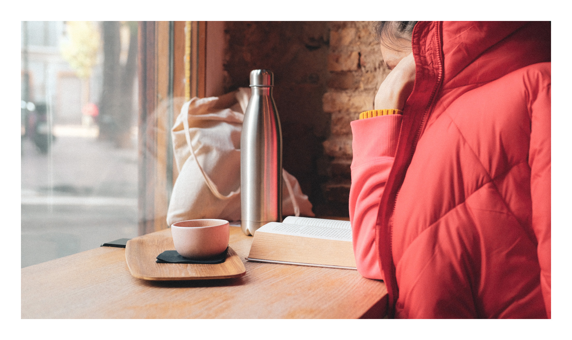 Placer II: lectura y café. Justicia & Coffee. Una chica leyendo en una cafetería, delante de una ventana que da a la calla, con su abrigo rojo por encima para taparse