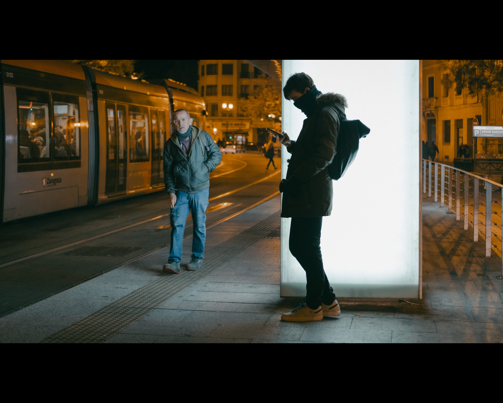 Esperando al tranvía a primera hora. Un chico, a primera hora de la mañana, espera al tranvía mirando el móvil y apoyado sobre la cristalera con luz artificial