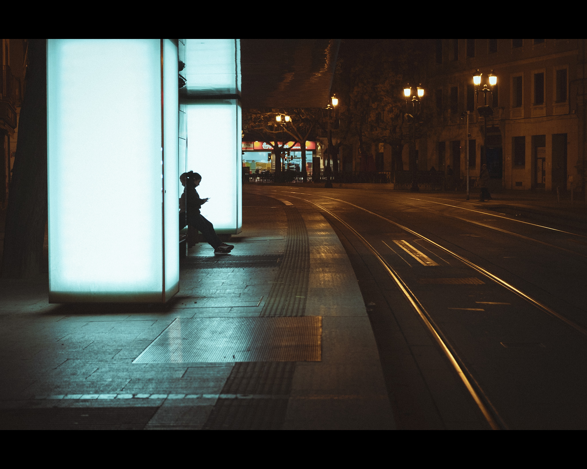 Una chica espera al tranvía mirando el móvil y se ve su figura a contraluz por la iluminación artificial de la parada del tranvía