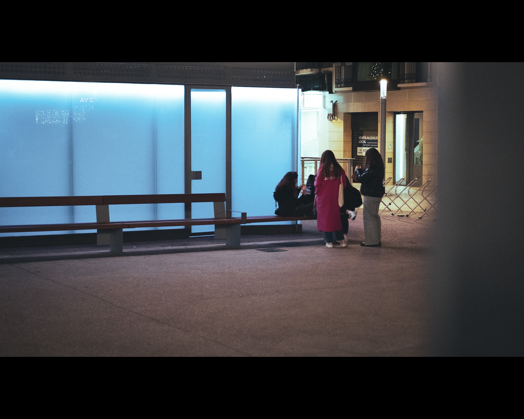 A la fresca navideña. Un grupo de chicas charlando al aire libre en una plaza con una pared de cristal iluminada con una luz intensa y azulada