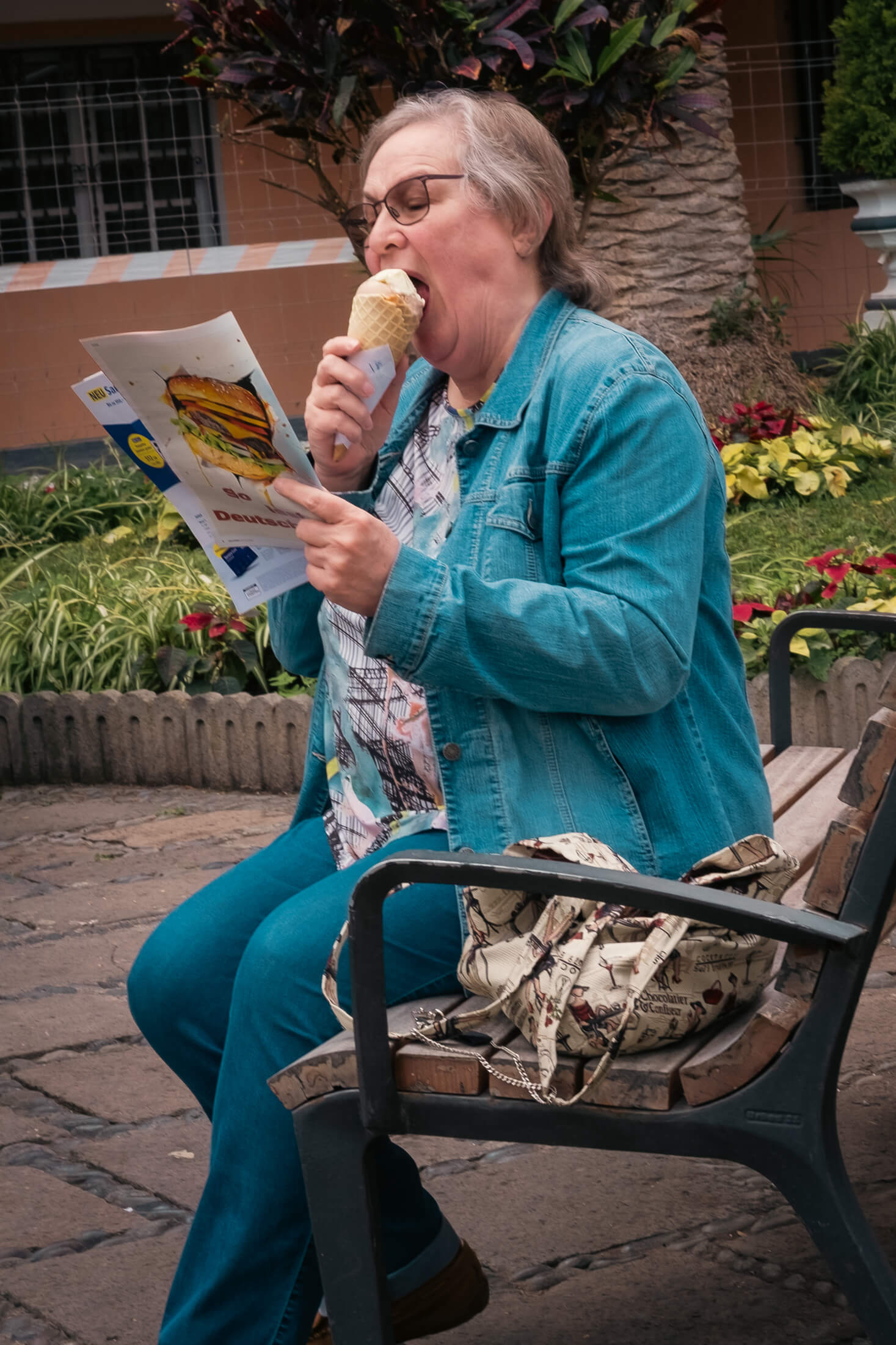 Foto de una señora leyendo una revista que tiene una foto de una hamburguesa mientras está dando un buen bocado al helado de cucurucho que sostiene con la otra mano