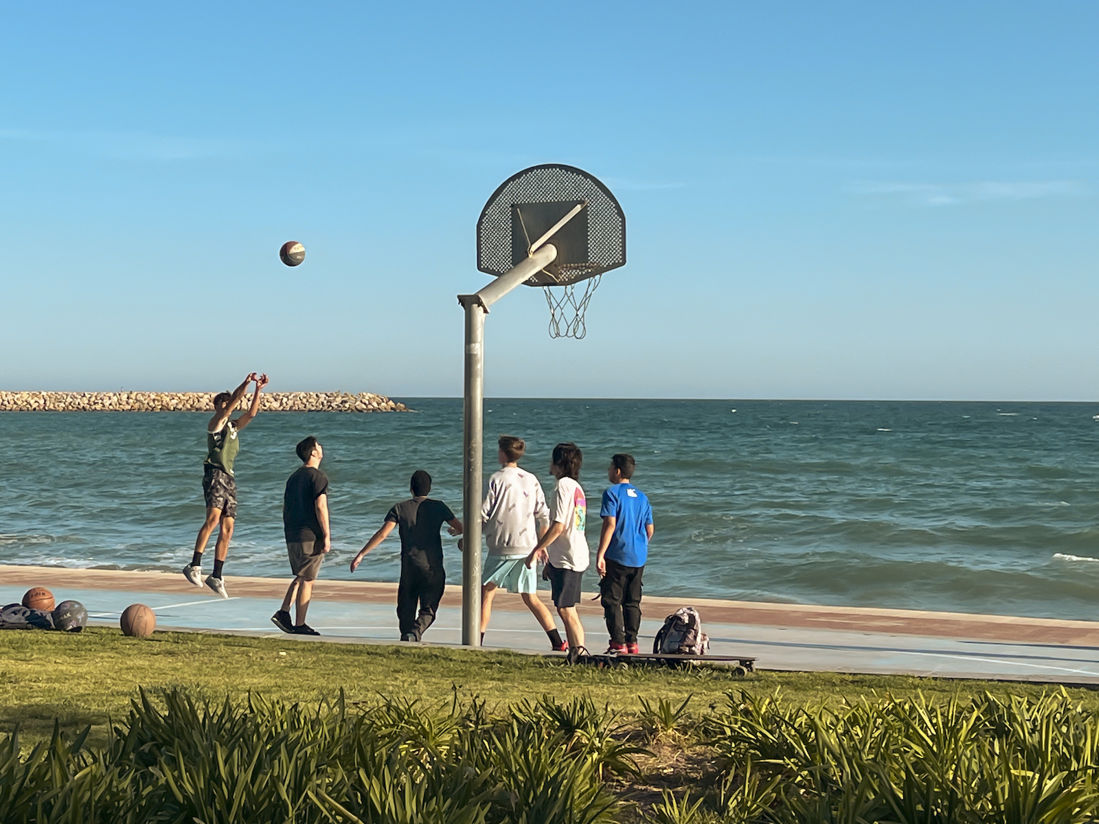Adolescentes jugando al baloncesto en la playa, justo en el momento de tirar un triple, con el balón en el aire.