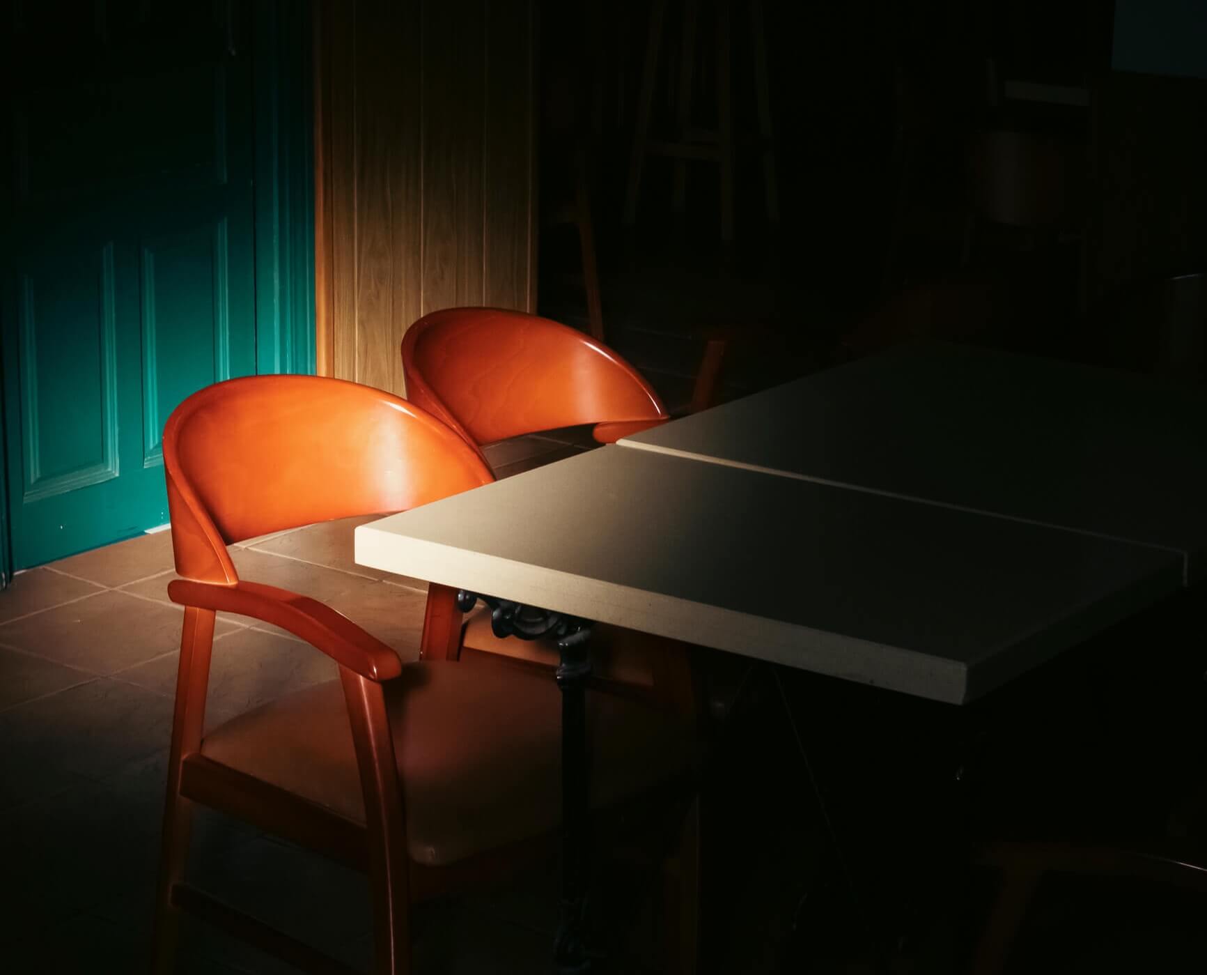 En un bar de hotel está muy oscuro salbo la zona que ilumina la luz del amanecer que empieza a entrar por una ventana y que ilumuna una silla y una mesa muy antiguas.