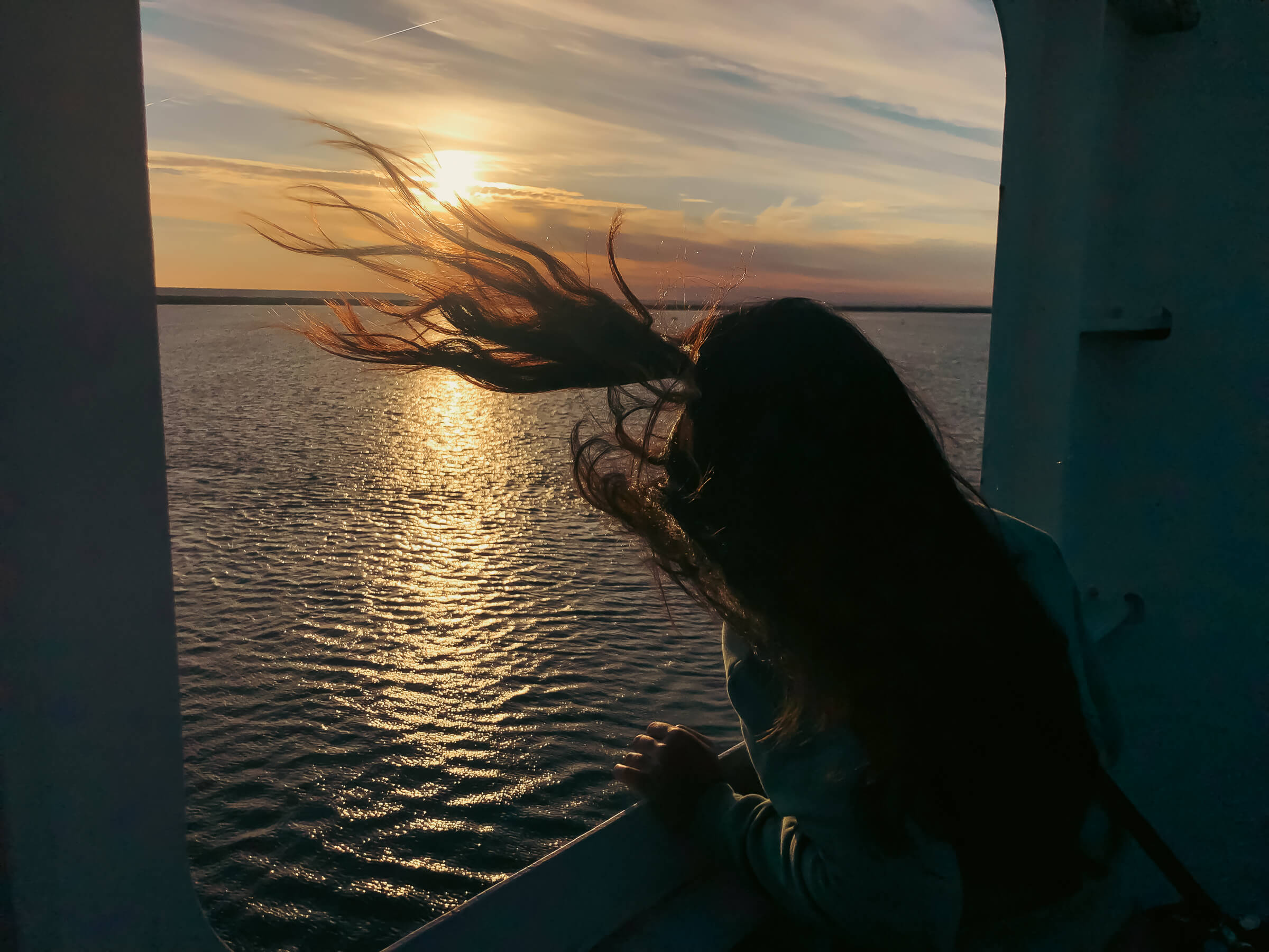 Laura viendo la llegada al puerto de Huelva al atardecer. Laura asomándose para ver la llegada al puerto de Huelva, con el sol poniéndose y los pelos moviéndose con el viento.
